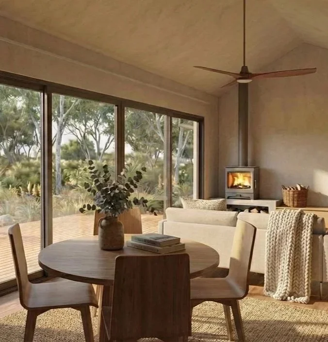 View of scope 1 bedroom retreat living area, with large double glazed windows, a round wooden dining table with books and wood fire, situated in a natural setting.