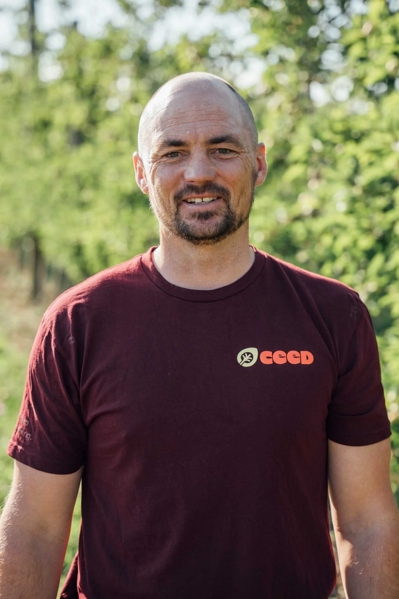 A man outdoors in a wooded area, wearing a maroon t-shirt with a logo that reads 'CEED'.