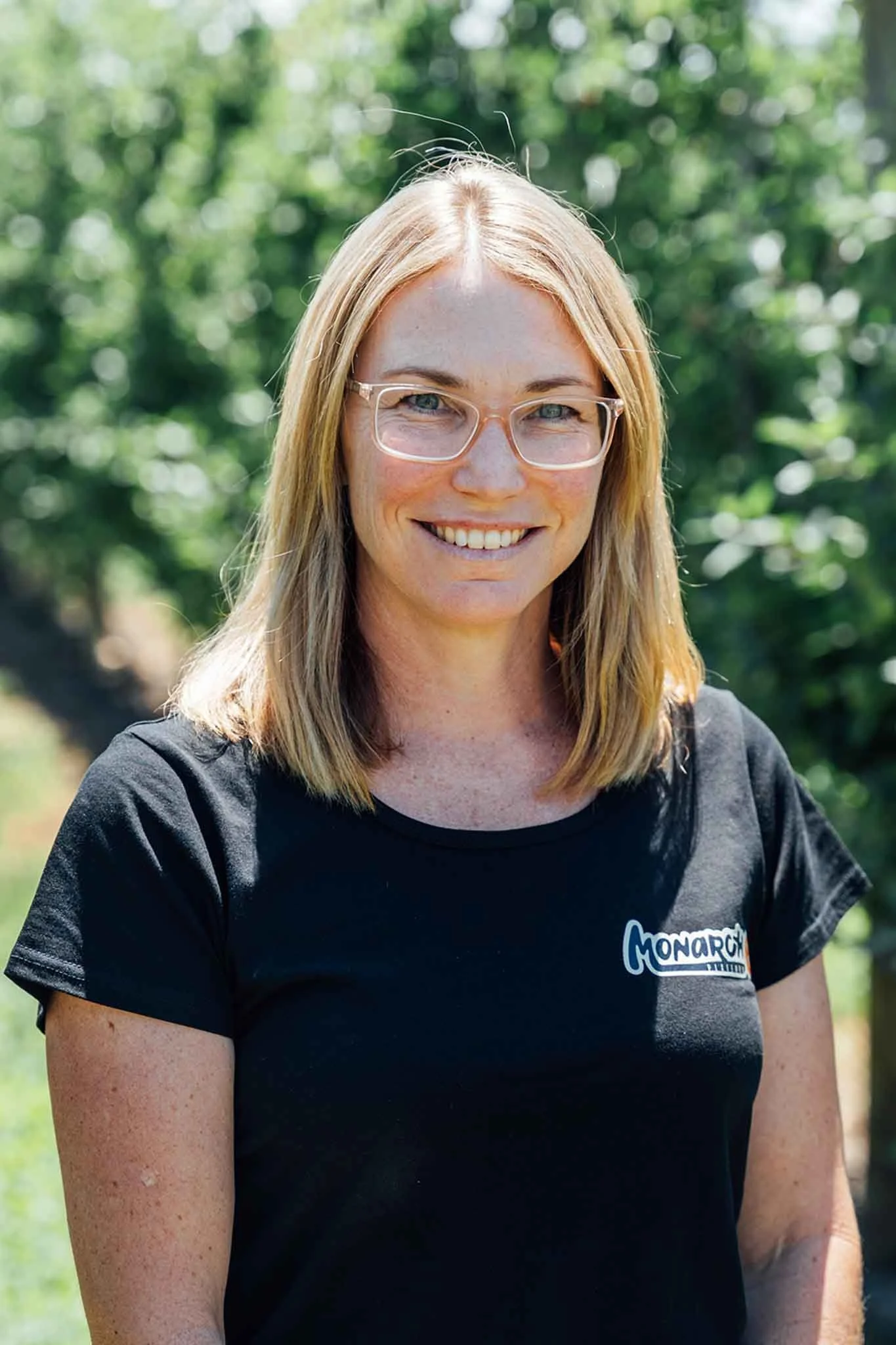 A woman with shoulder-length blonde hair, glasses, and a black T-shirt with a logo, standing outdoors in a park with green trees and sunlight.