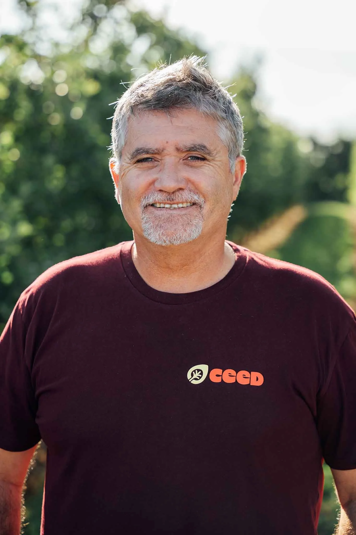 A smiling middle-aged man with gray hair and a beard, wearing a maroon T-shirt with 