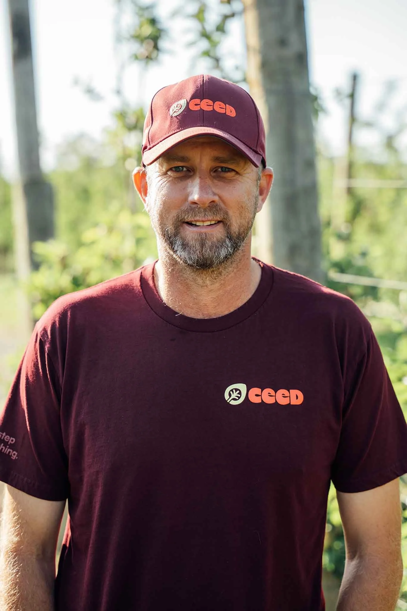 A man wearing a maroon cap and maroon T-shirt with 'CEED' logo, standing outdoors with trees and a partly cloudy sky in the background.