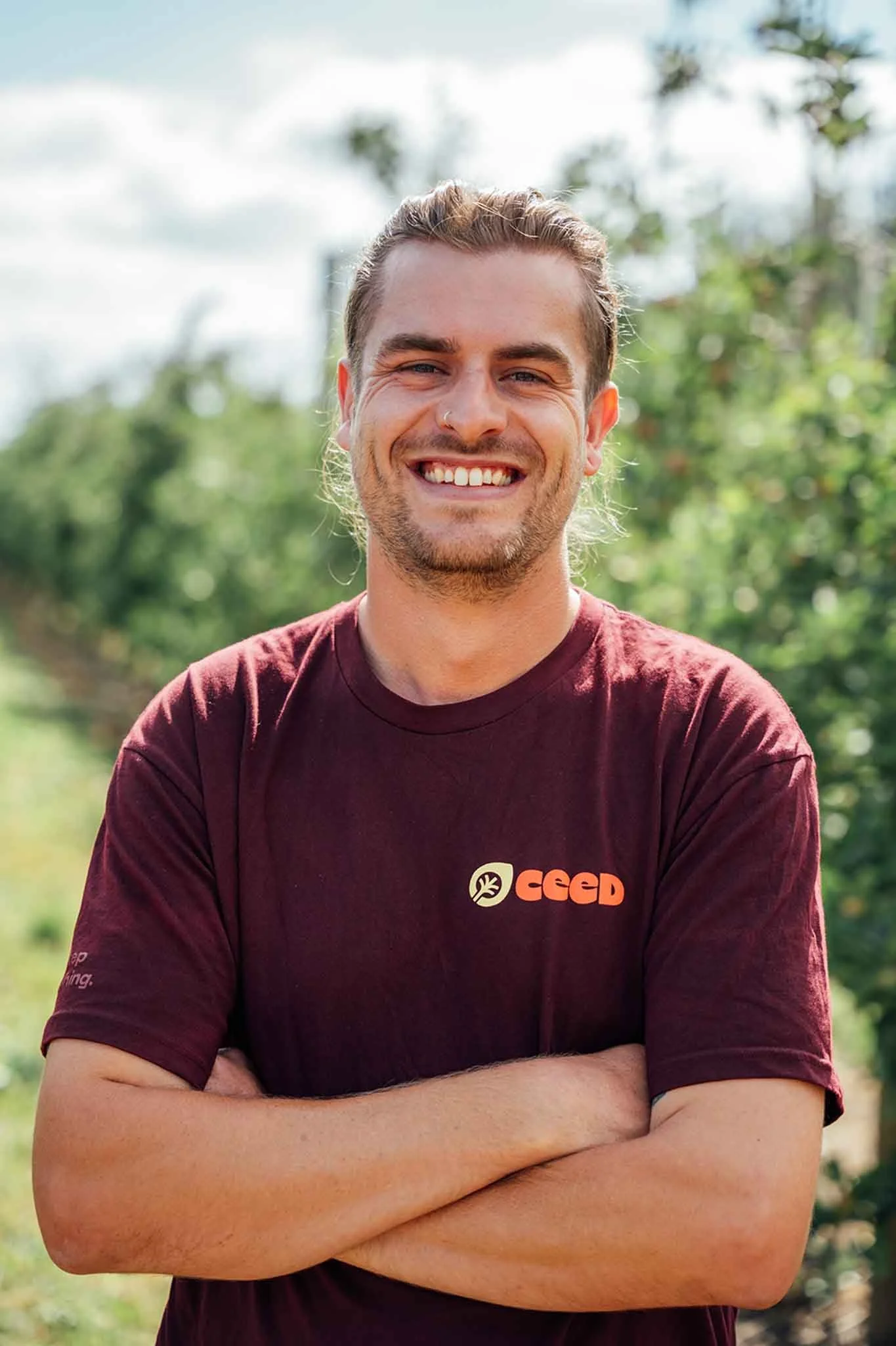 A young man with a smile, light brown hair, wearing a maroon t-shirt with a logo on it, standing outdoors with green trees and a partly cloudy sky in the background.