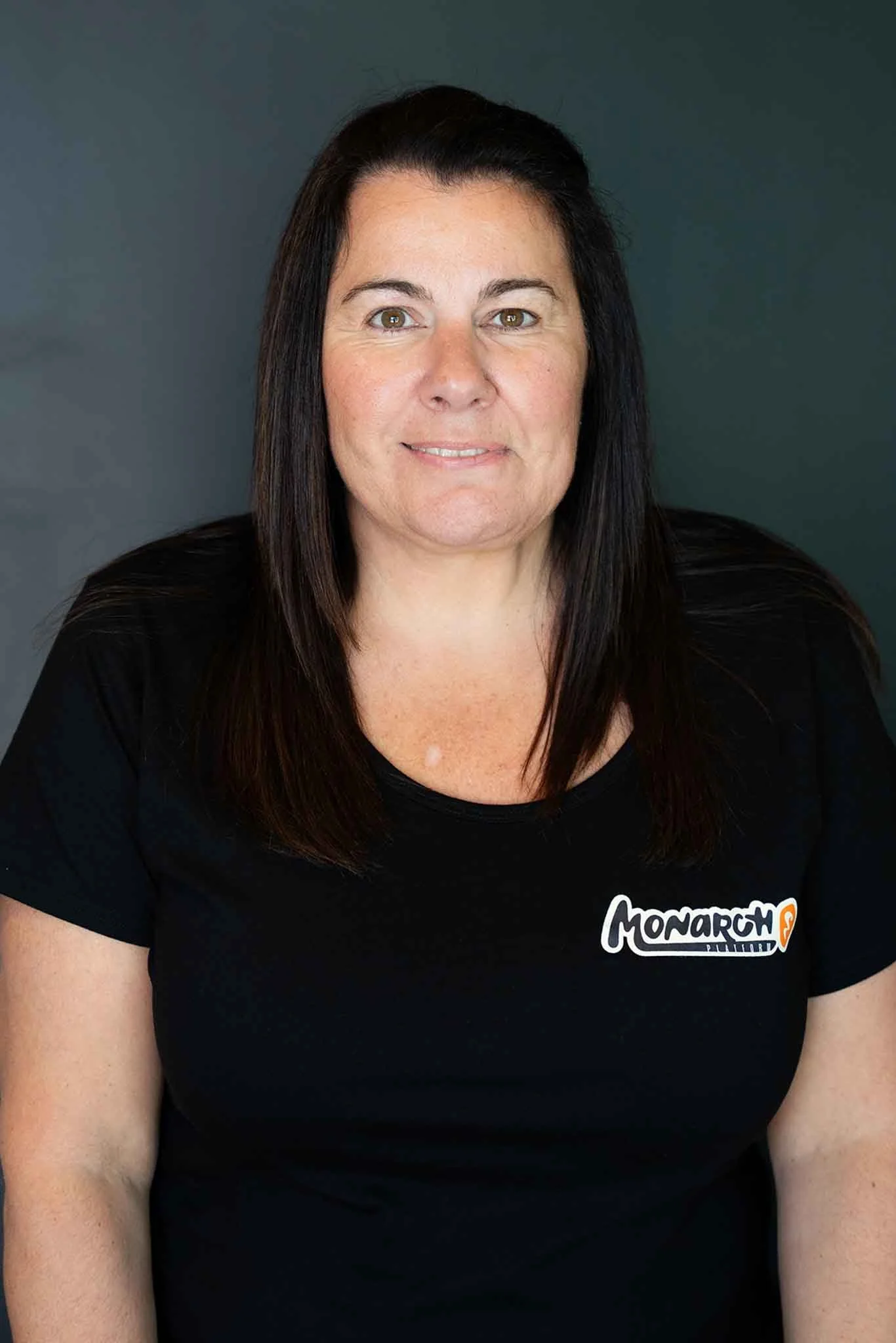 A woman with dark brown hair wearing a black t-shirt with a logo that says 'Monarch' in white and orange, against a dark green background.