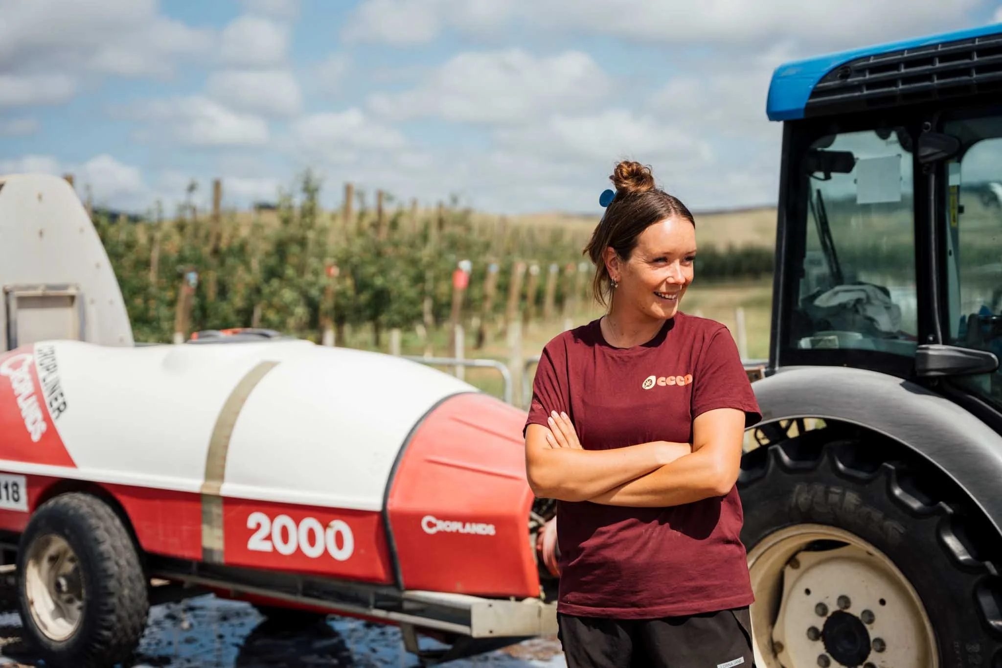 A woman standing outdoors near a large piece of farming or agricultural equipment, smiling and wearing a maroon T-shirt, on a sunny day with trees and blue sky in the background.