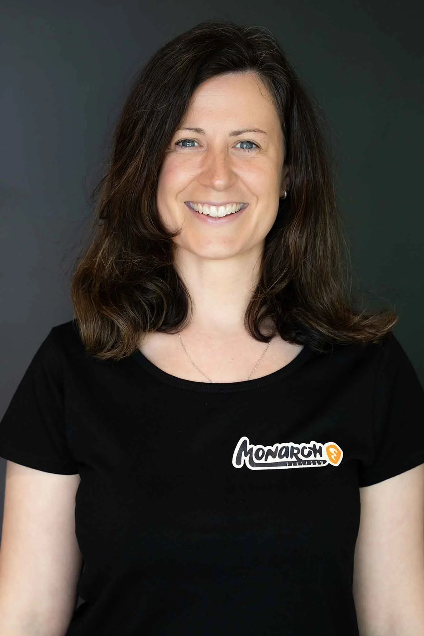 A woman with shoulder-length brown hair, smiling, wearing a black t-shirt with a Monarch logo on it, against a dark background.