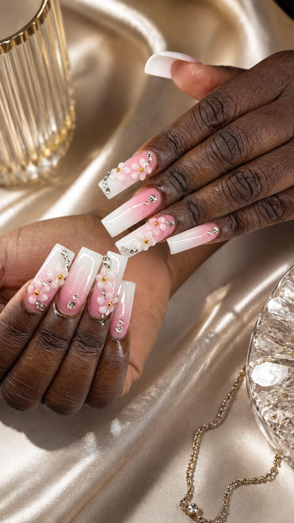 Close-up of two hands with elaborate pink and white acrylic nails featuring floral and jewel embellishments, resting on a satin fabric surface with jewelry nearby.