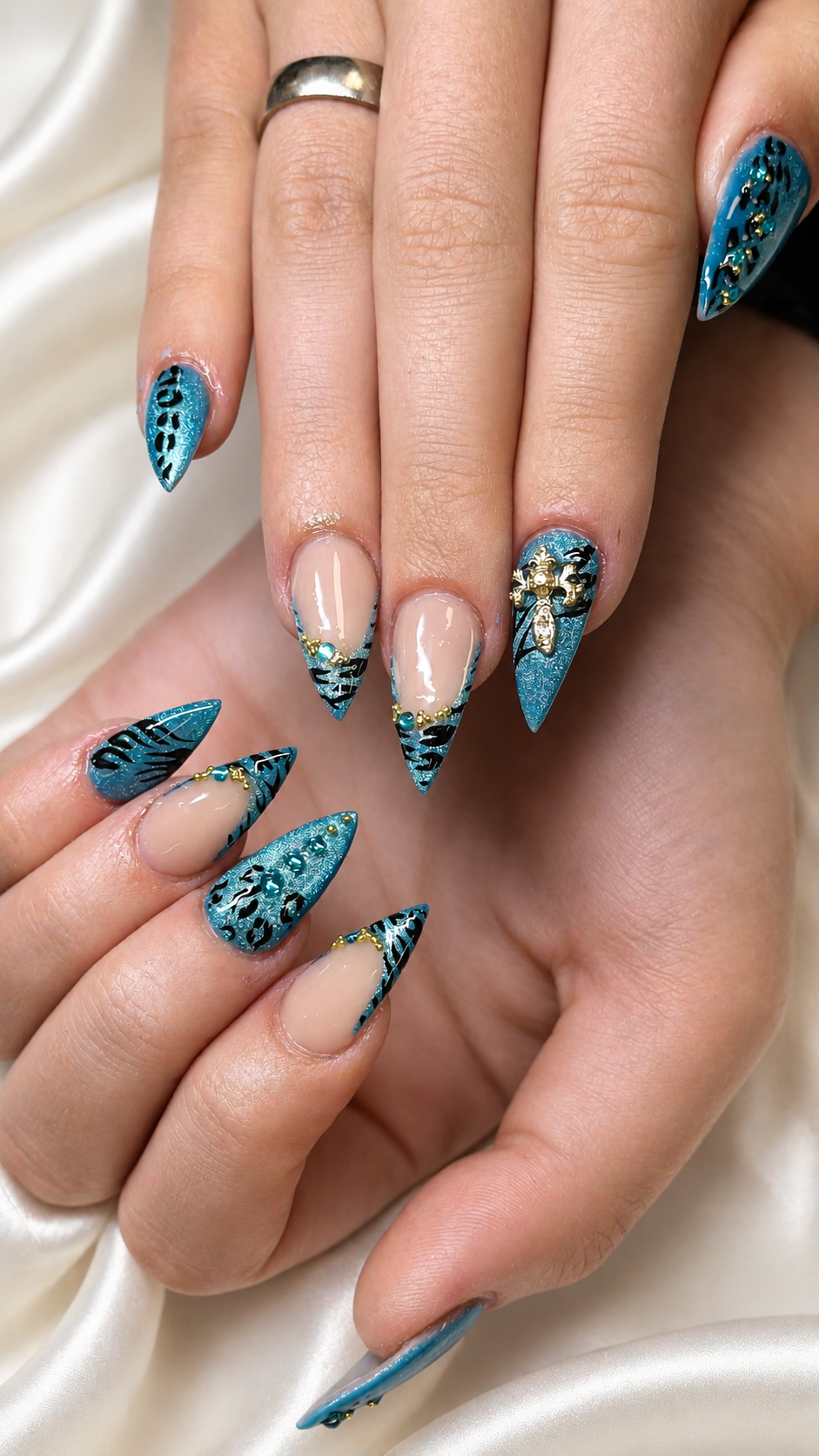 Close-up of hands with long, pointed nails painted in blue and black tiger stripe pattern, decorated with gold beads and small metallic accents. Some nails have intricate designs, including a small floral embellishment.