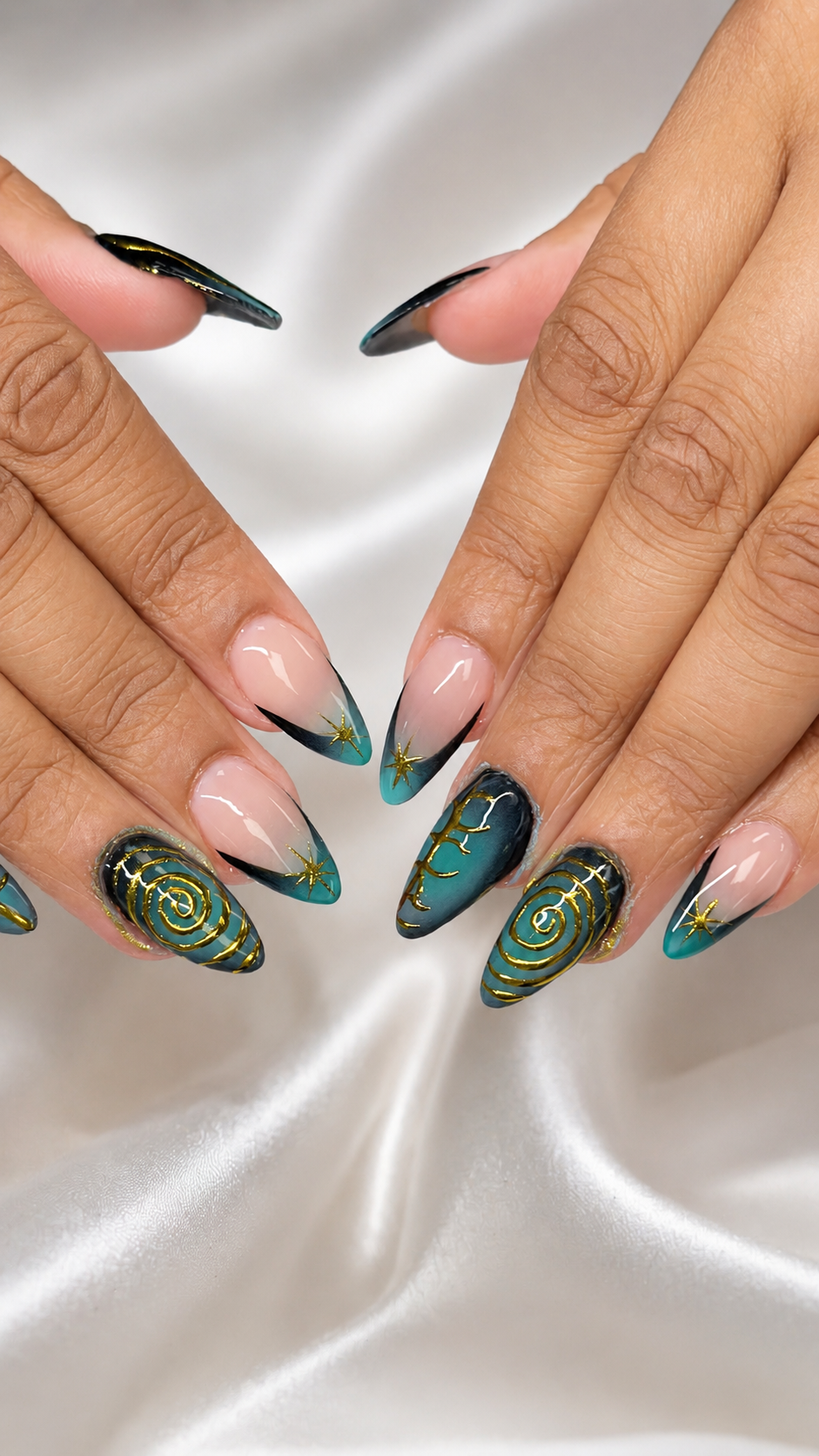 Nail art with black, teal, and gold spiral and star designs on long coffin-shaped nails, set against a satin white background.