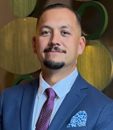 A man dressed in a blue suit with a light blue shirt and a purple tie, standing indoors with a background of large decorative green and yellow circles.