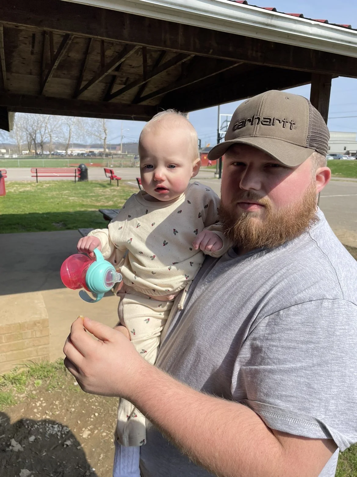 A man with a beard and a cap holding a young child with a sippy cup under a wooden shelter at a park.