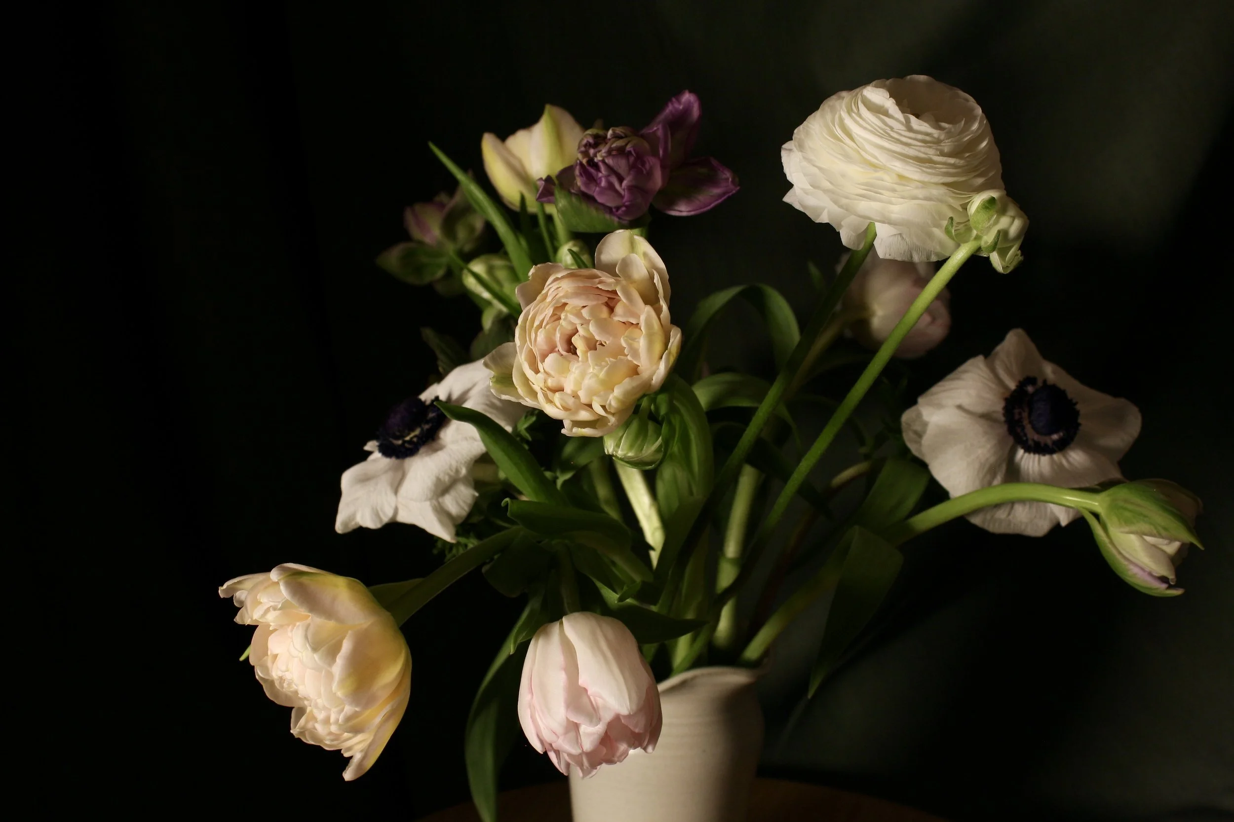 A bouquet of various tulips and ranunculus in white, pink, and purple in a beige vase against a dark background.