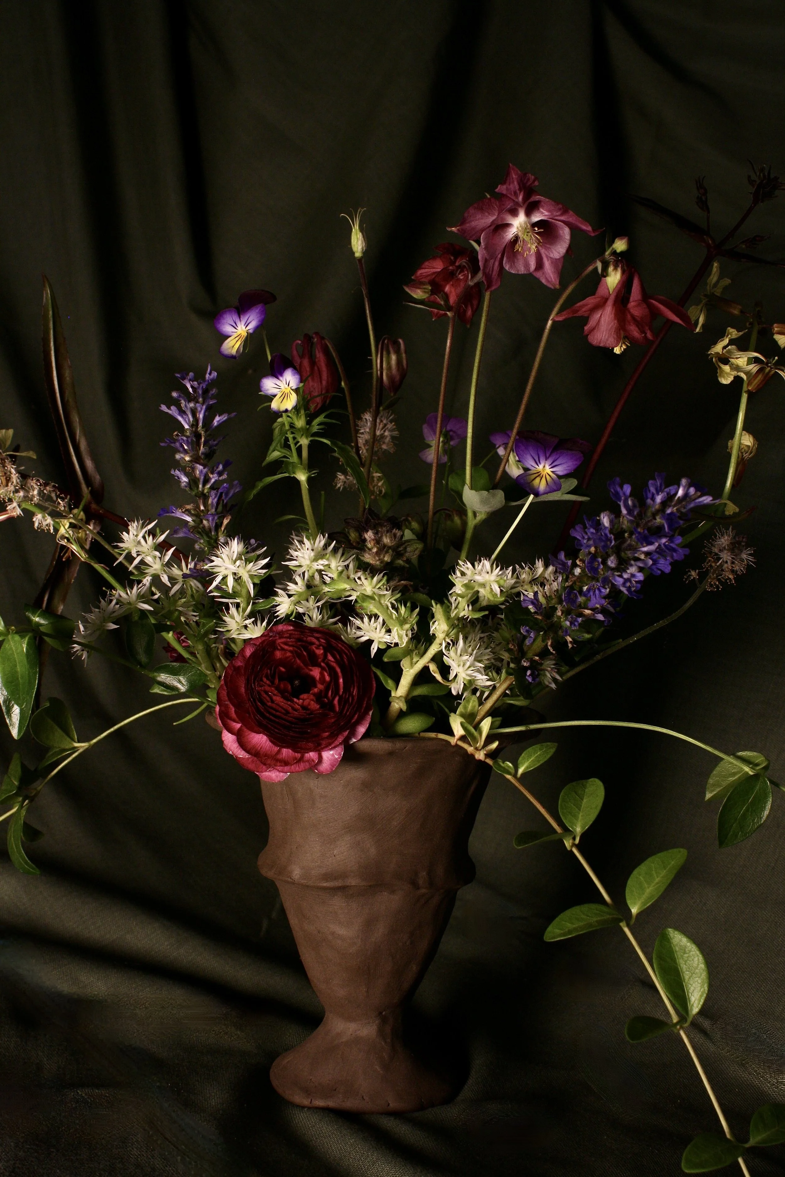A floral arrangement in a brown vase with purple, red, and white flowers and green foliage against a dark background.