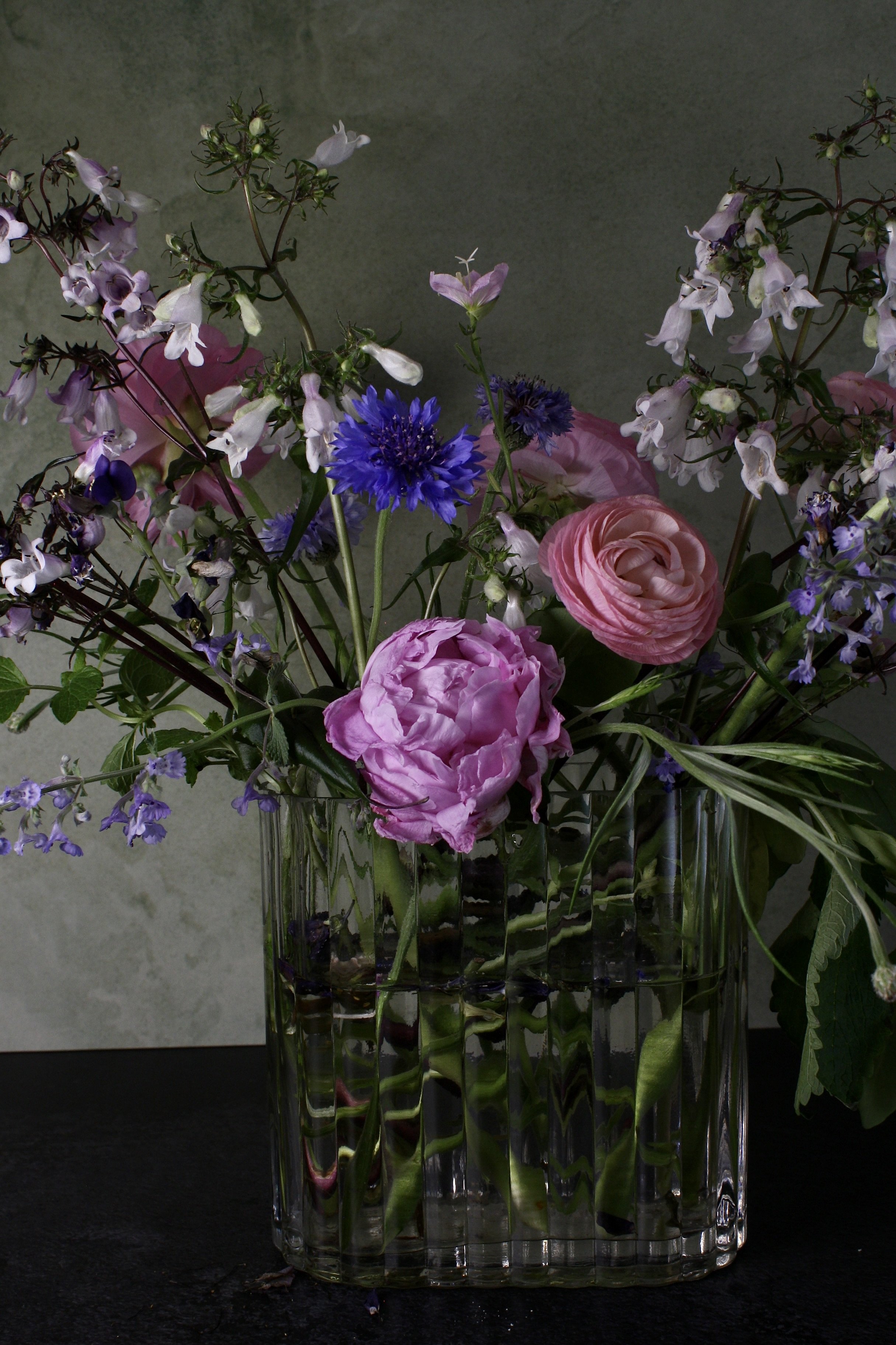 A bouquet of pink roses, purple cornflowers, and small white and purple blossoms in a clear, textured glass vase on a dark surface with a plain gray background.