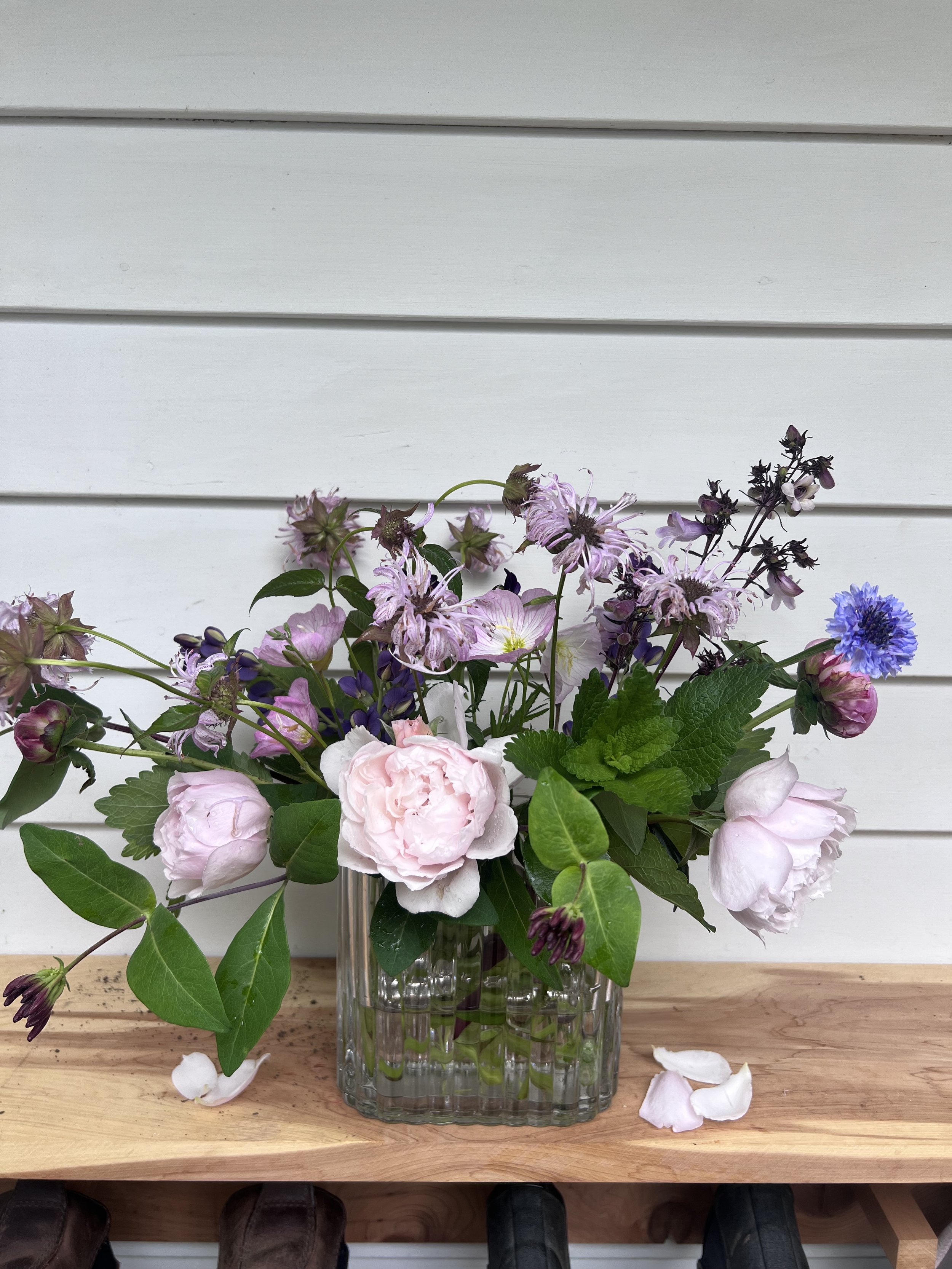 A floral arrangement in a clear ribbed glass vase on a wooden surface, with pink, purple, and blue flowers and green leaves, set against a white wooden background.