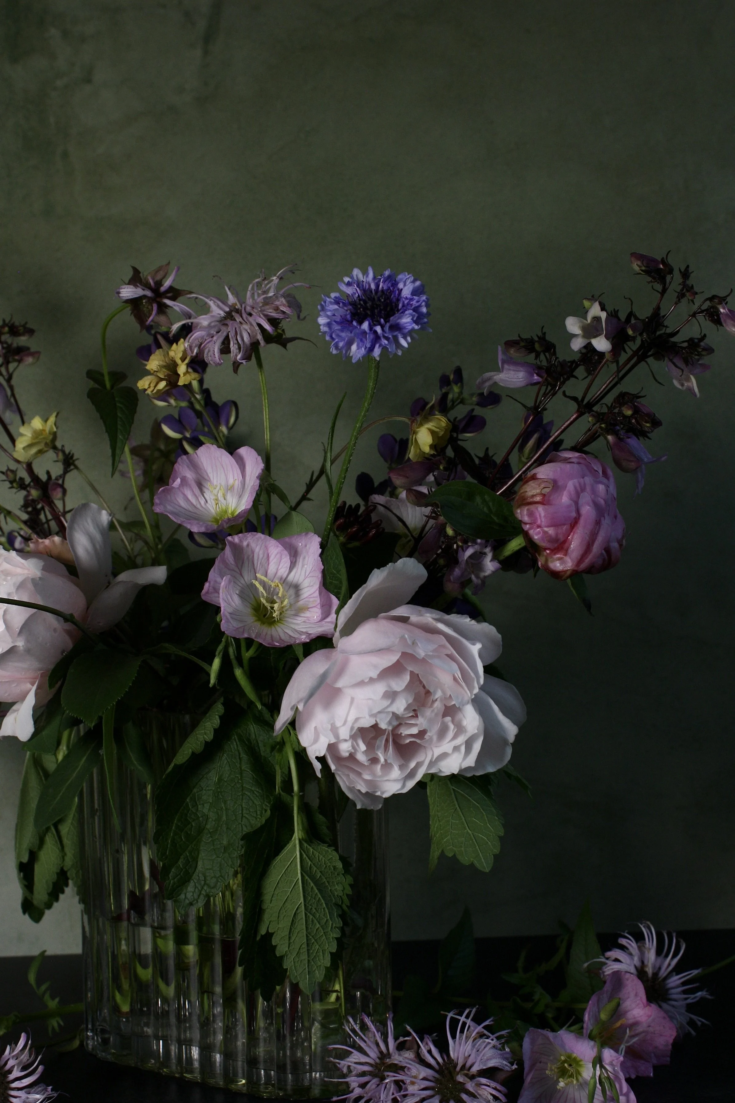 A floral arrangement in a tall glass vase featuring various pink, purple, white, and yellow flowers against a dark background.