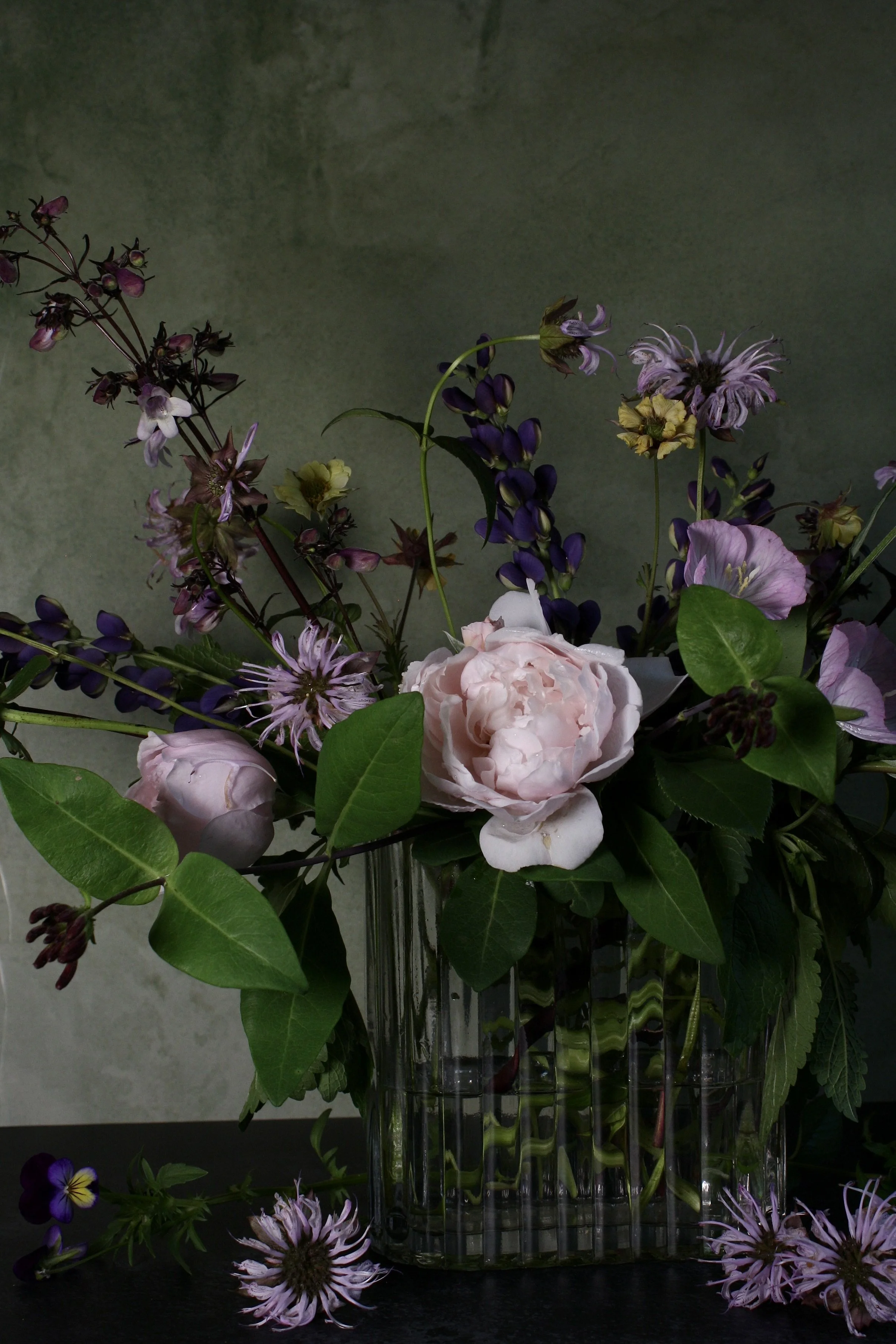 A bouquet of pink, purple, yellow, and white flowers arranged in a tall glass vase, with some flowers spilling onto a dark surface.