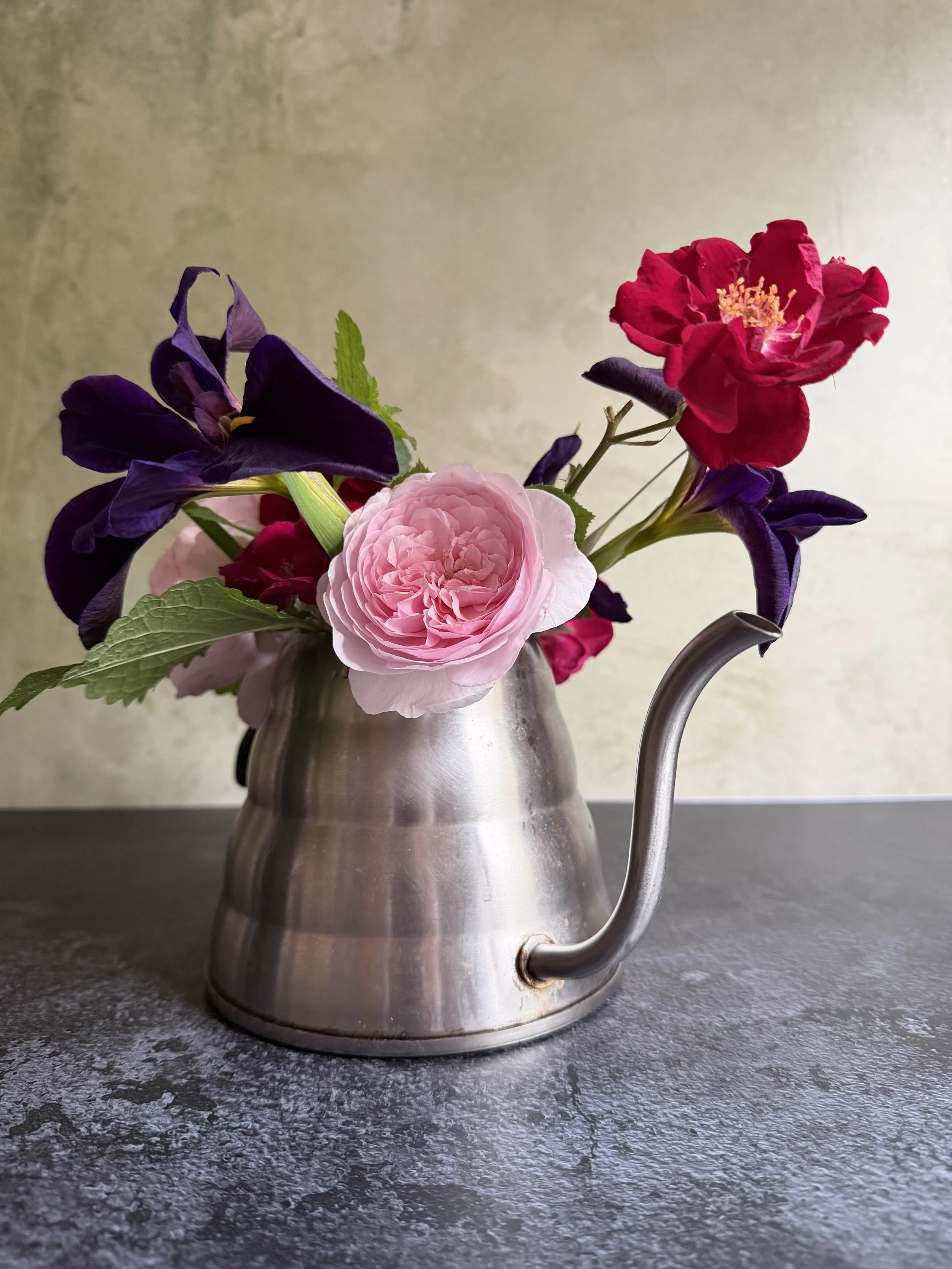 A metal watering can with a bouquet of purple, pink, and red flowers, placed on a textured dark gray surface against a plain light background.