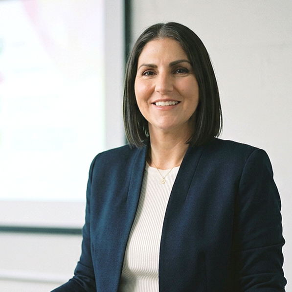 A woman with shoulder-length dark hair smiling, wearing a navy blazer and a white top, seated in a professional setting with a blurred presentation screen in the background.