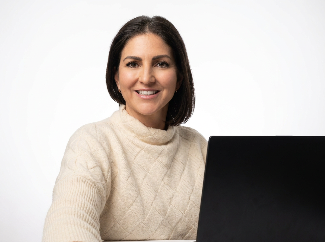 A woman with shoulder-length dark hair wearing a cream-colored sweater, sitting in front of a laptop, smiling at the camera against a white background.