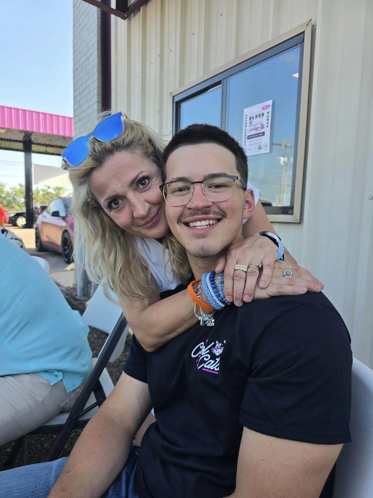 A woman and a young man are sitting outdoors, smiling, with the woman hugging the man from behind. The woman has blonde hair, blue sunglasses on her head, and is wearing jewelry. The man has short dark hair, glasses, and is wearing a black t-shirt wi