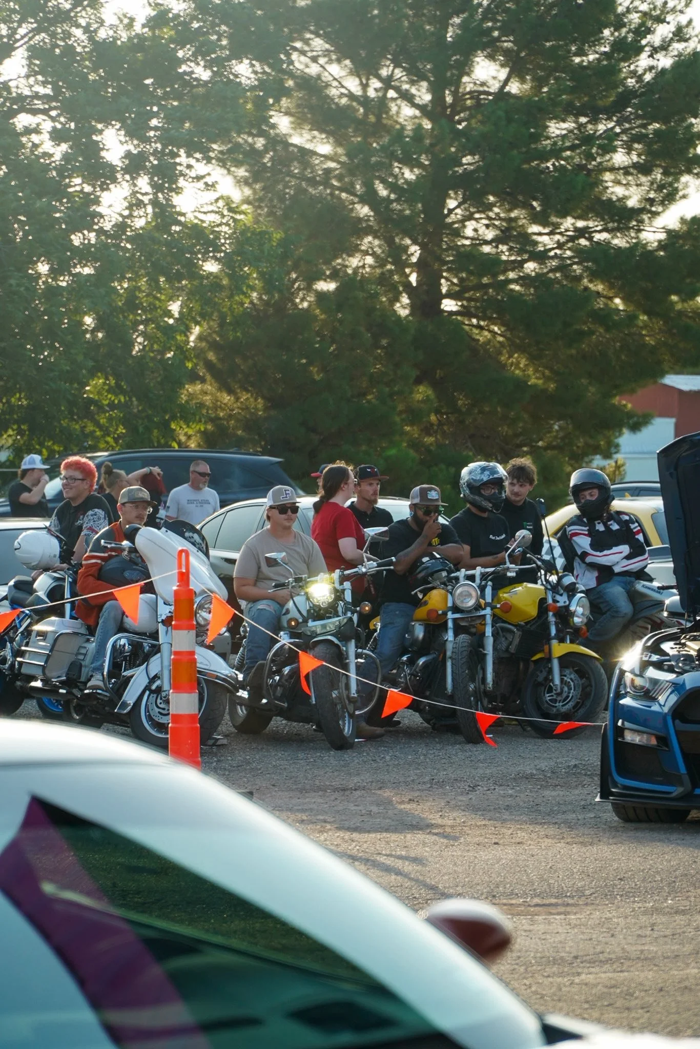 Group of people, some wearing helmets, gathered with motorcycles and cars in an outdoor parking lot, with large trees and a sunset in the background.