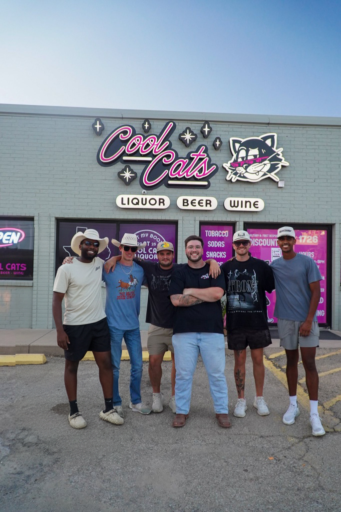 Group of six young men standing in front of a store called 'Cool Cats' with neon sign, cigarettes, liquor, beer, and wine listed, during daytime.