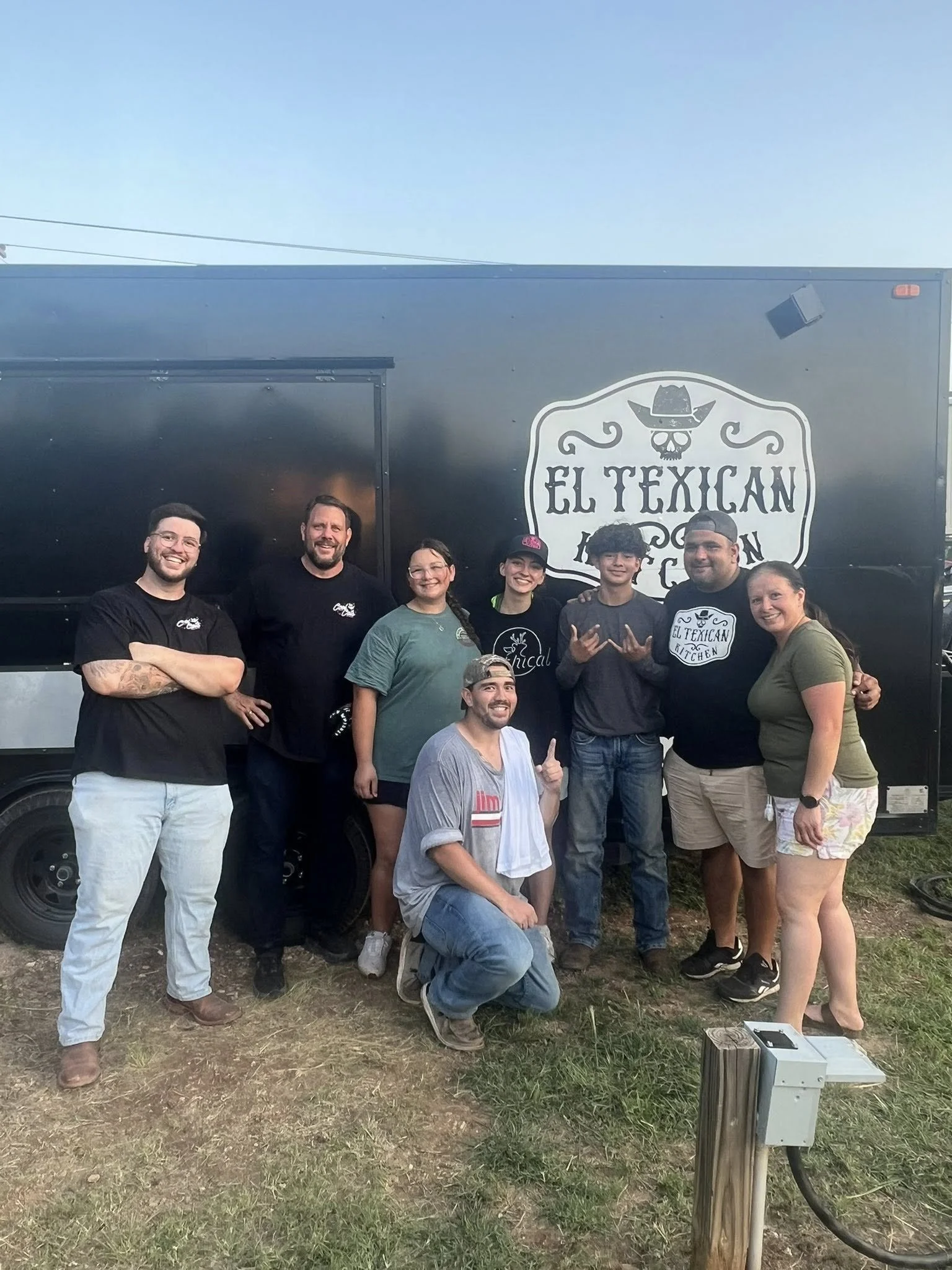 Group of seven people standing in front of a black food truck with a sign that reads "El Texican Kitchen" and a graphic of a skull wearing a cowboy hat. They are smiling and posing for the photo outdoors.