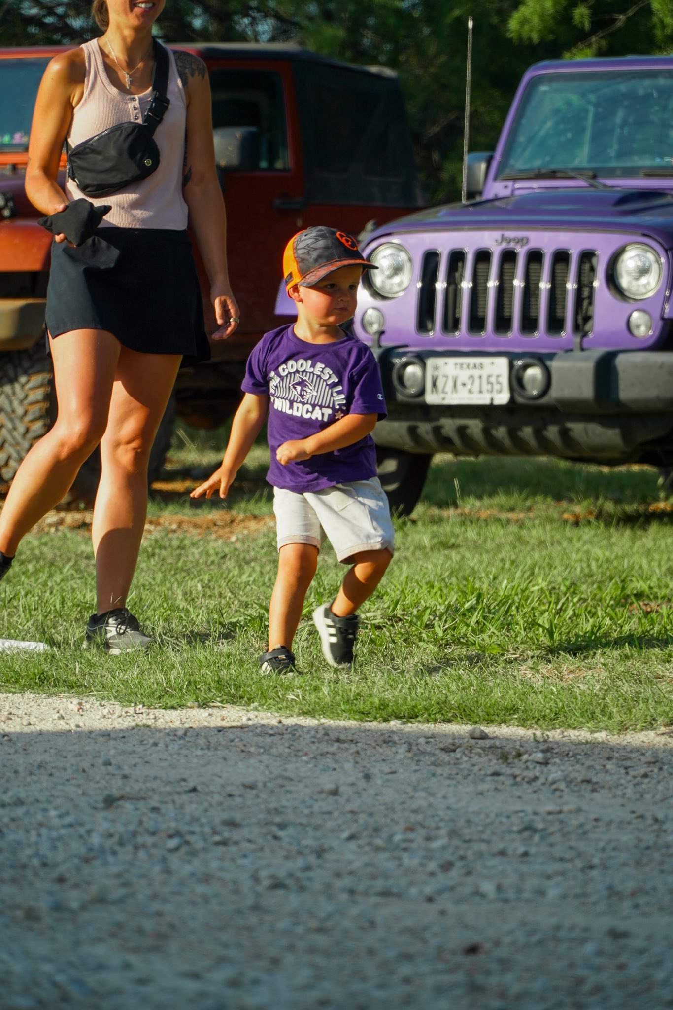 A young boy in a purple t-shirt and shorts playing on grass with a woman nearby and colorful vehicles in the background.