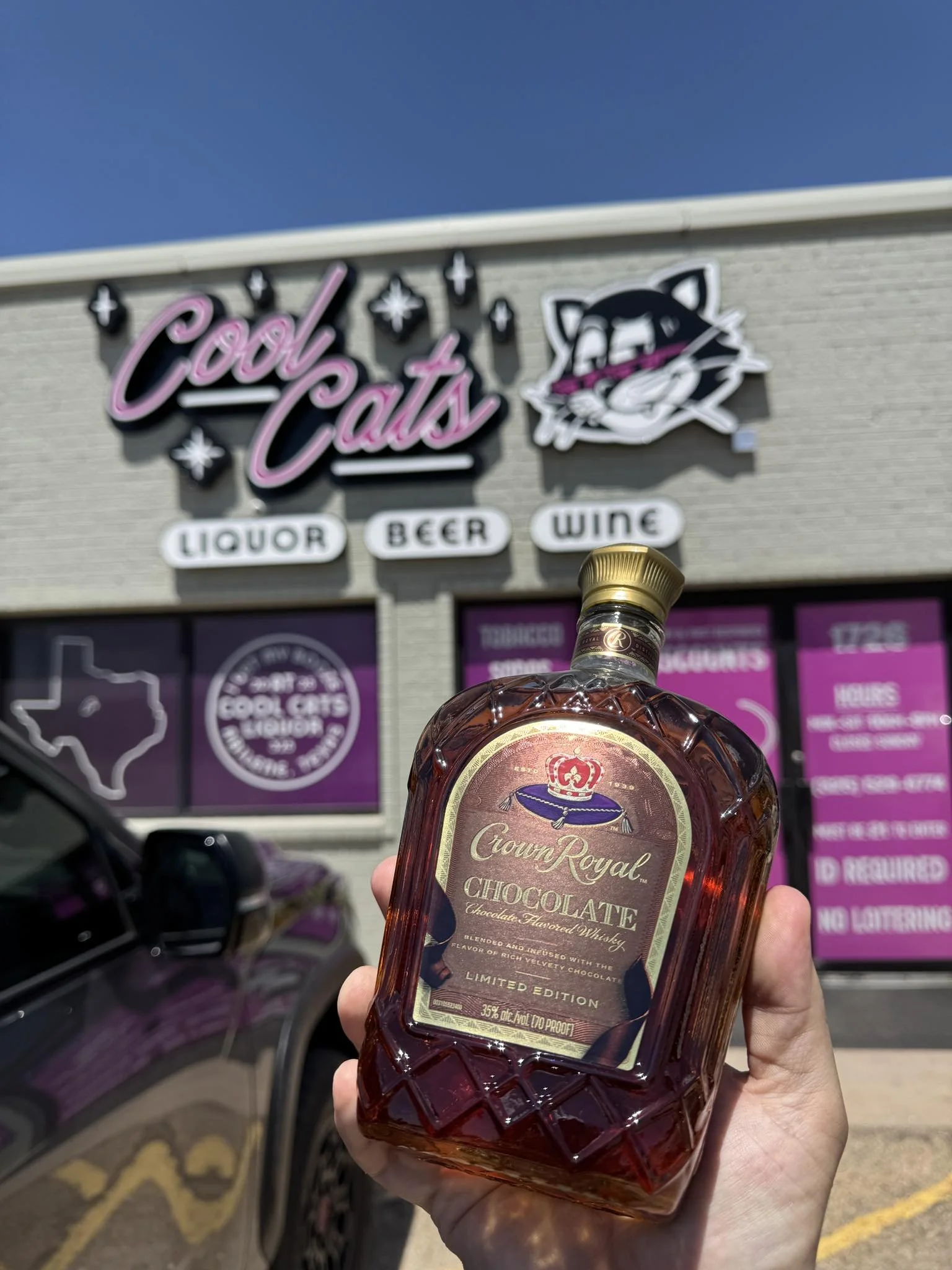 A person holding a bottle of Crown Royal Chocolate whiskey outside a store called 'Cool Cats' that sells liquor, beer, and wine. The store has a pink and black sign with a cat logo and a map of Texas on the window.
