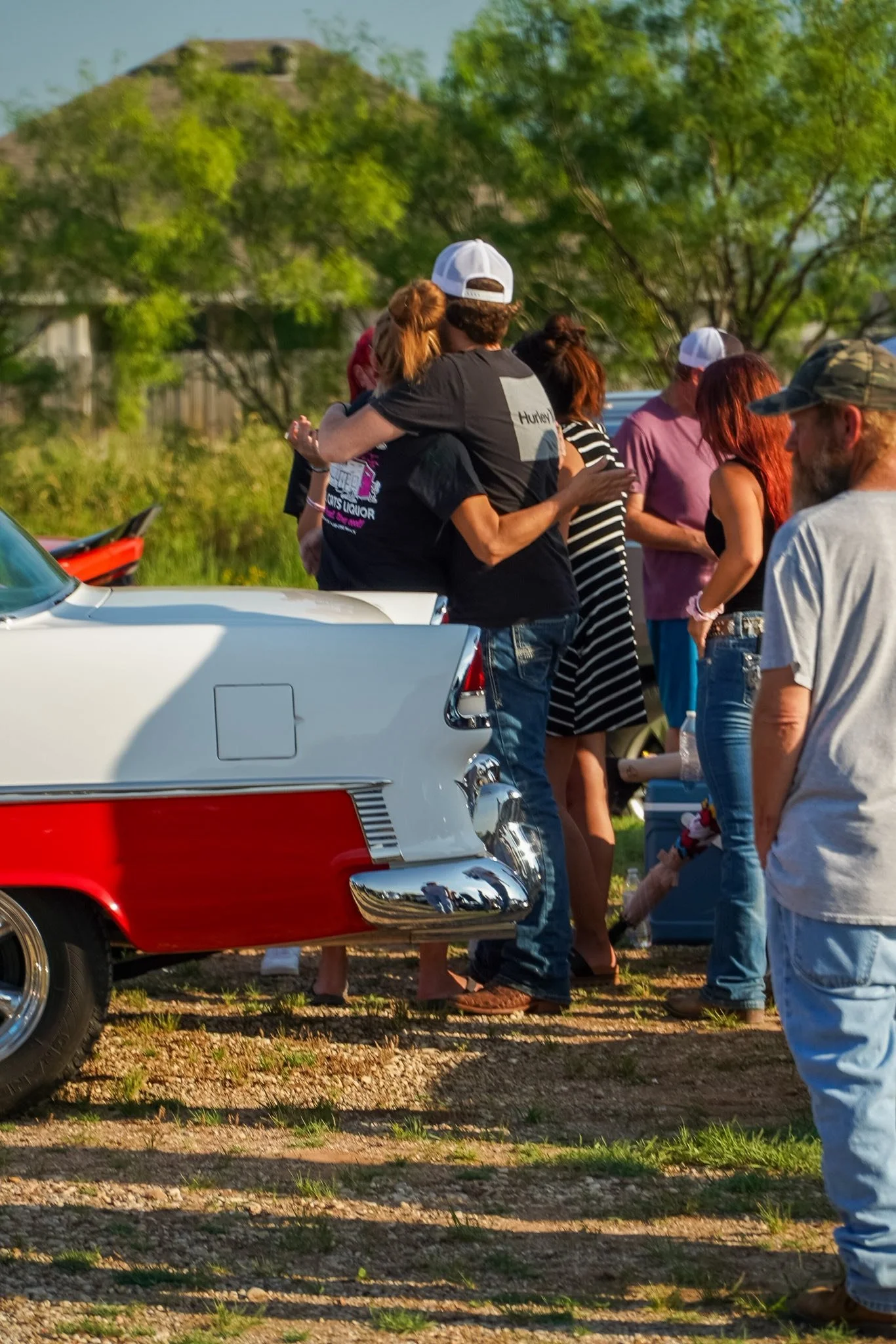A group of people gathering outdoors near vintage cars, with two people hugging, and others chatting, in a sunny setting with trees and a grassy area.