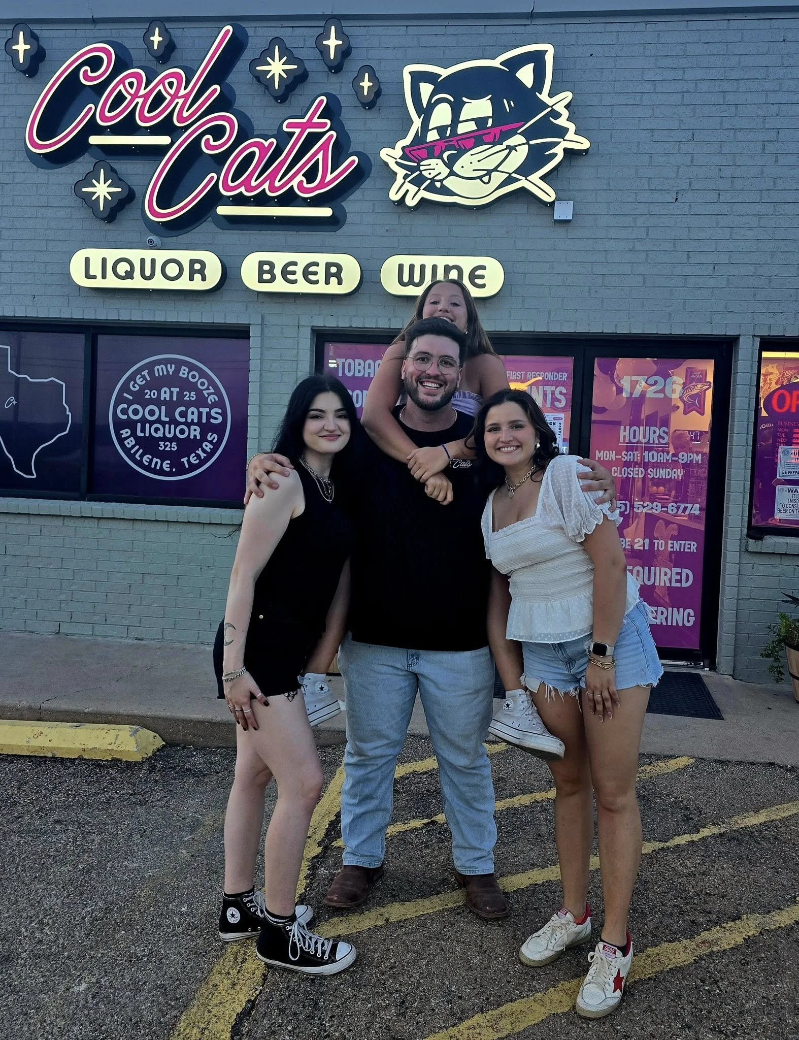 Four friends standing outside a liquor store called 'Cool Cats'. The store has a neon sign with a cat face and signs advertising liquor, beer, and wine. The group is smiling, with one person sitting on another's shoulders and others standing beside h