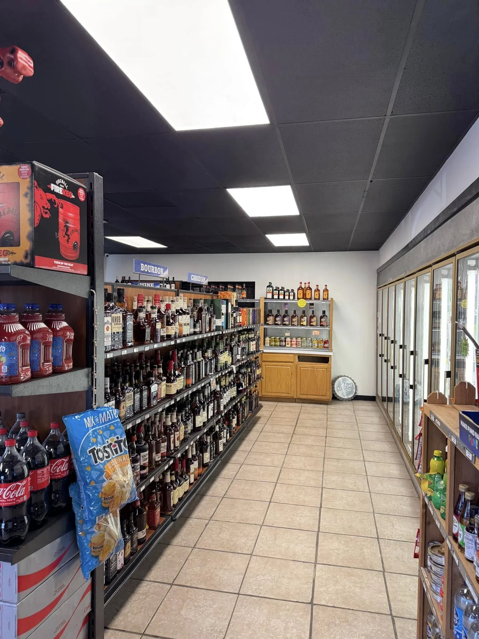 Shelves stocked with various bottles of alcoholic beverages in a liquor store aisle, with a refrigerated section on the right and signs for bourbon and Canadian whiskey in the background.