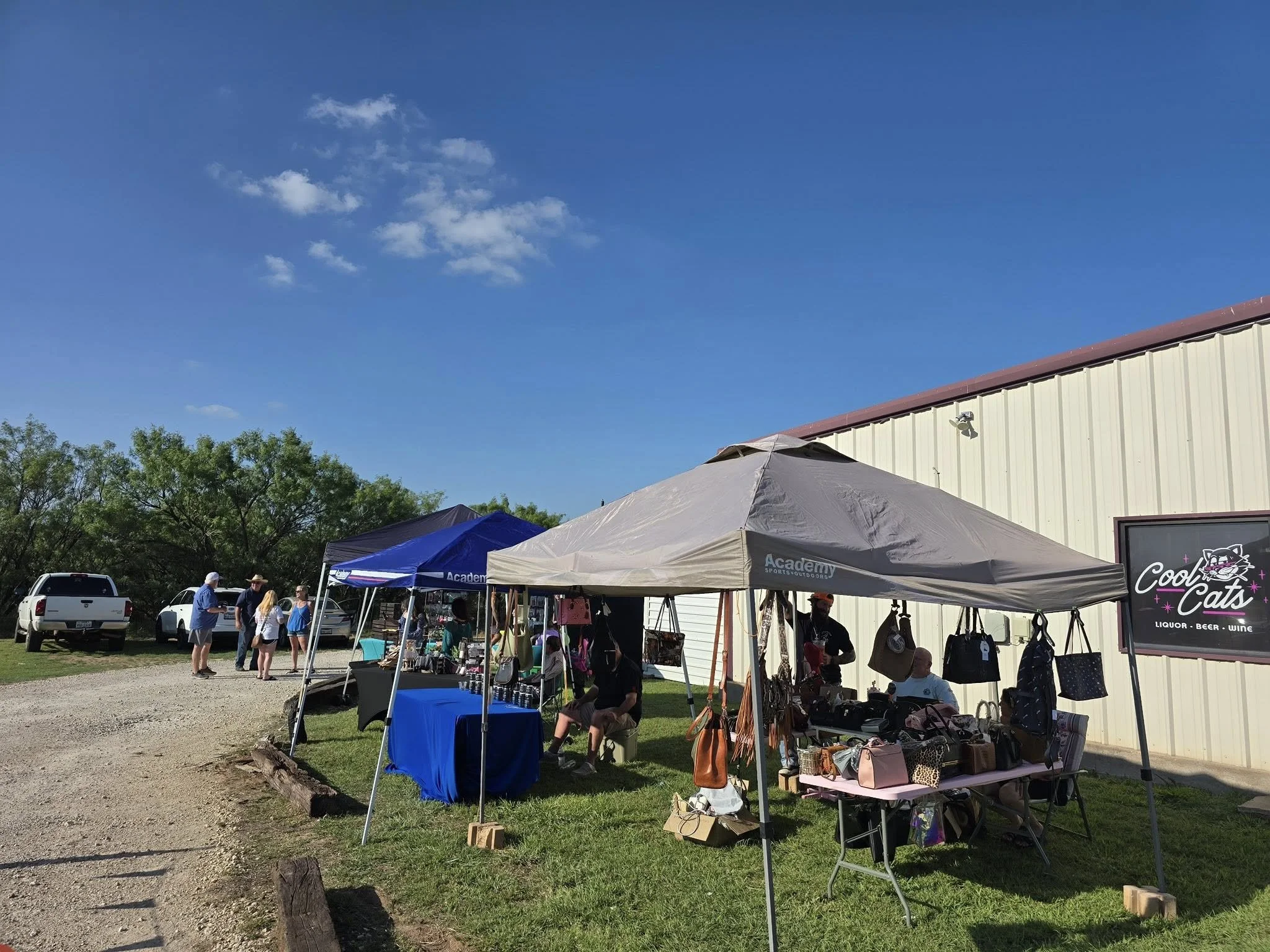 Outdoor market scene with tents, vendors, and shoppers on a sunny day, next to a light-colored building with a sign that says 'Cool Cats'.