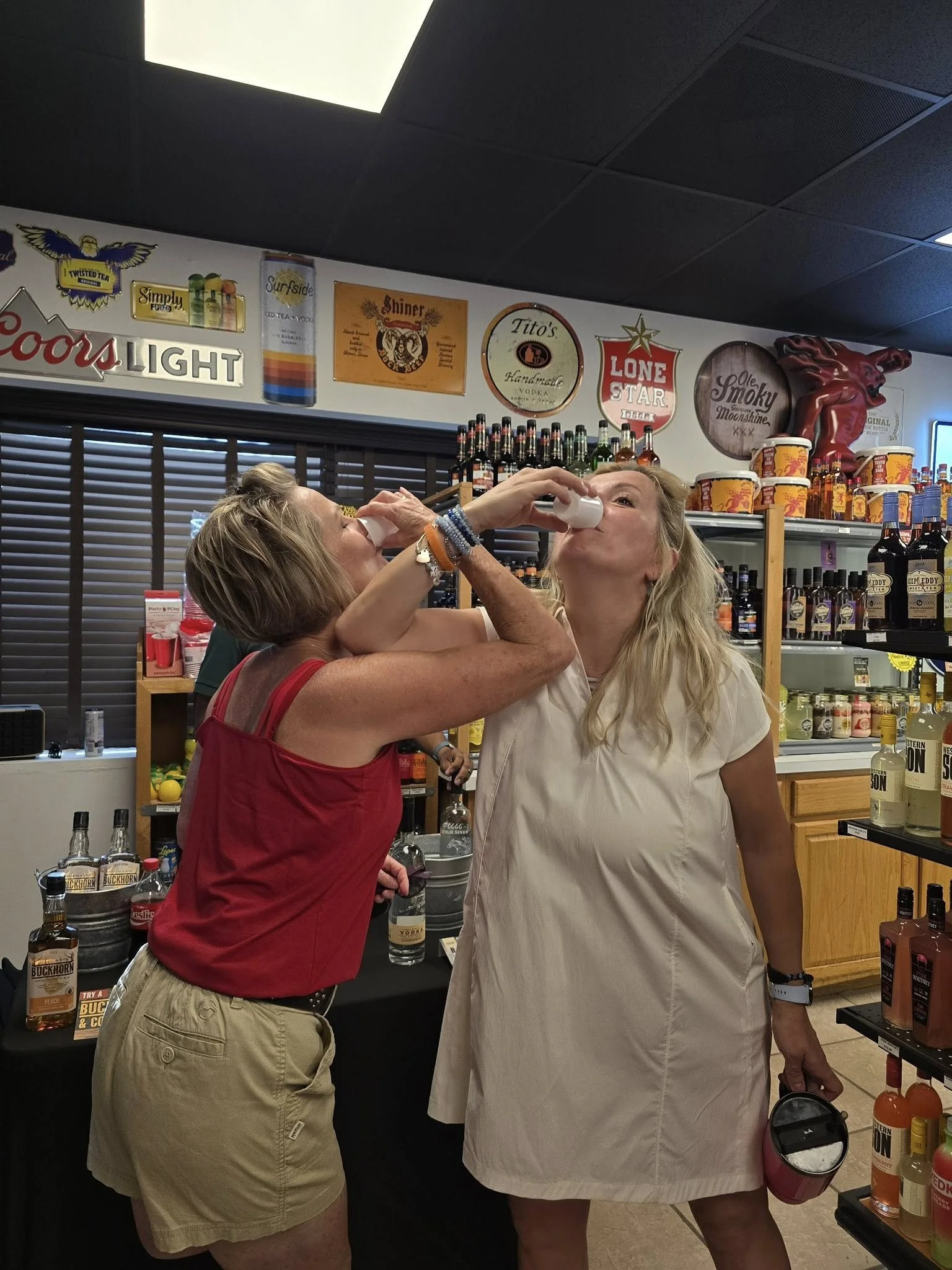 Two women are at a liquor store, one handing the other a shot of alcohol, with shelves of bottles behind them.