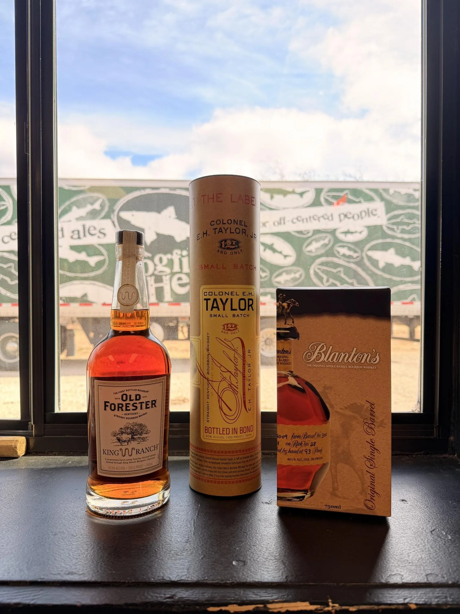 A bottle of Old Forester bourbon whiskey, a cylindrical container of Colonel E. H. Taylor bourbon, and a box of Blanton's bourbon whiskey on a black surface with a window showing a truck and trees outside.