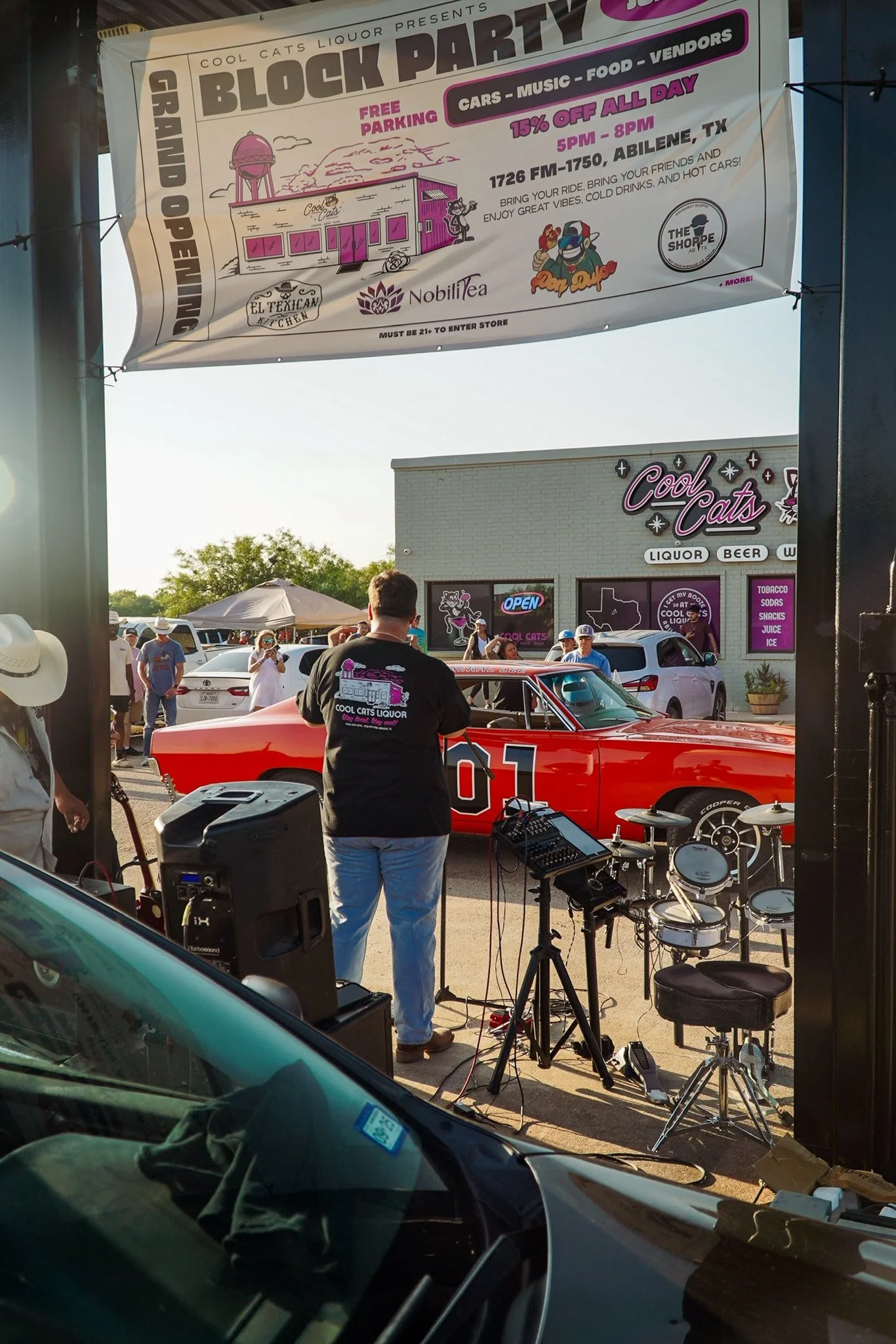A man standing in front of a red race car with the number 01, setup with musical instruments and equipment, during a block party event outside a store named Cool Cats Liquor, with a crowd and tents in the background, and a large sign for the event hanging overhead.
