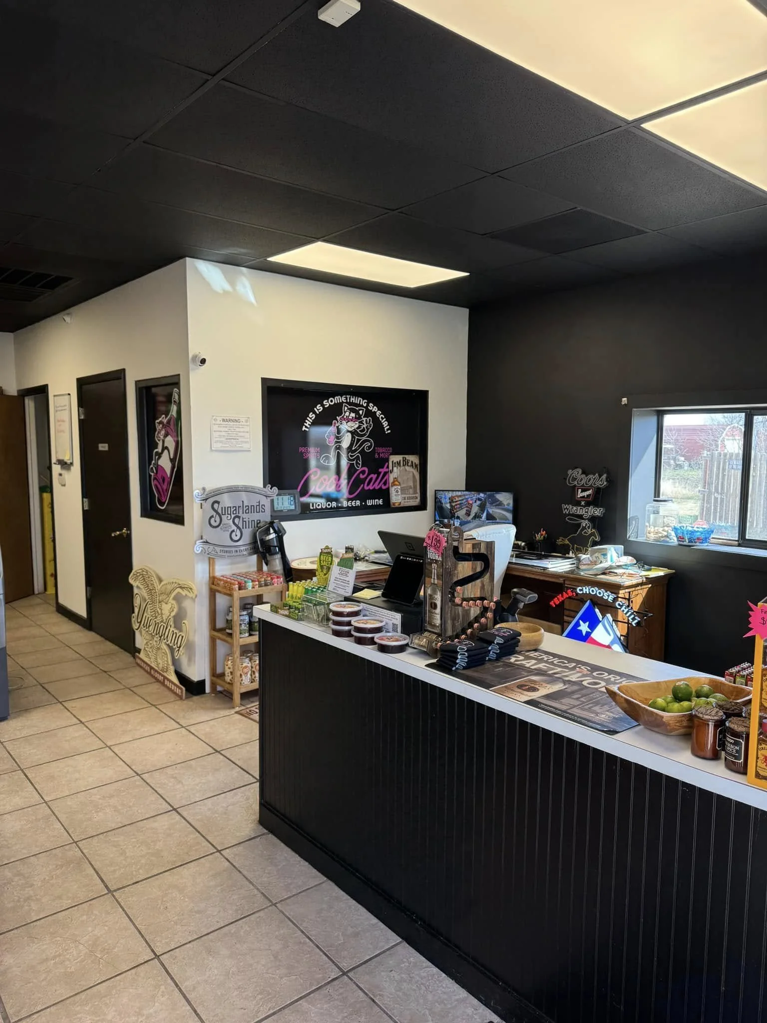 Interior of a small shop or cafe with a counter, various signs, and products on display including jars, lime, and snacks. The counter has a black front panel, and there's a window behind. The walls are decorated with signs and artwork, and ceiling tiles are black with fluorescent lighting.