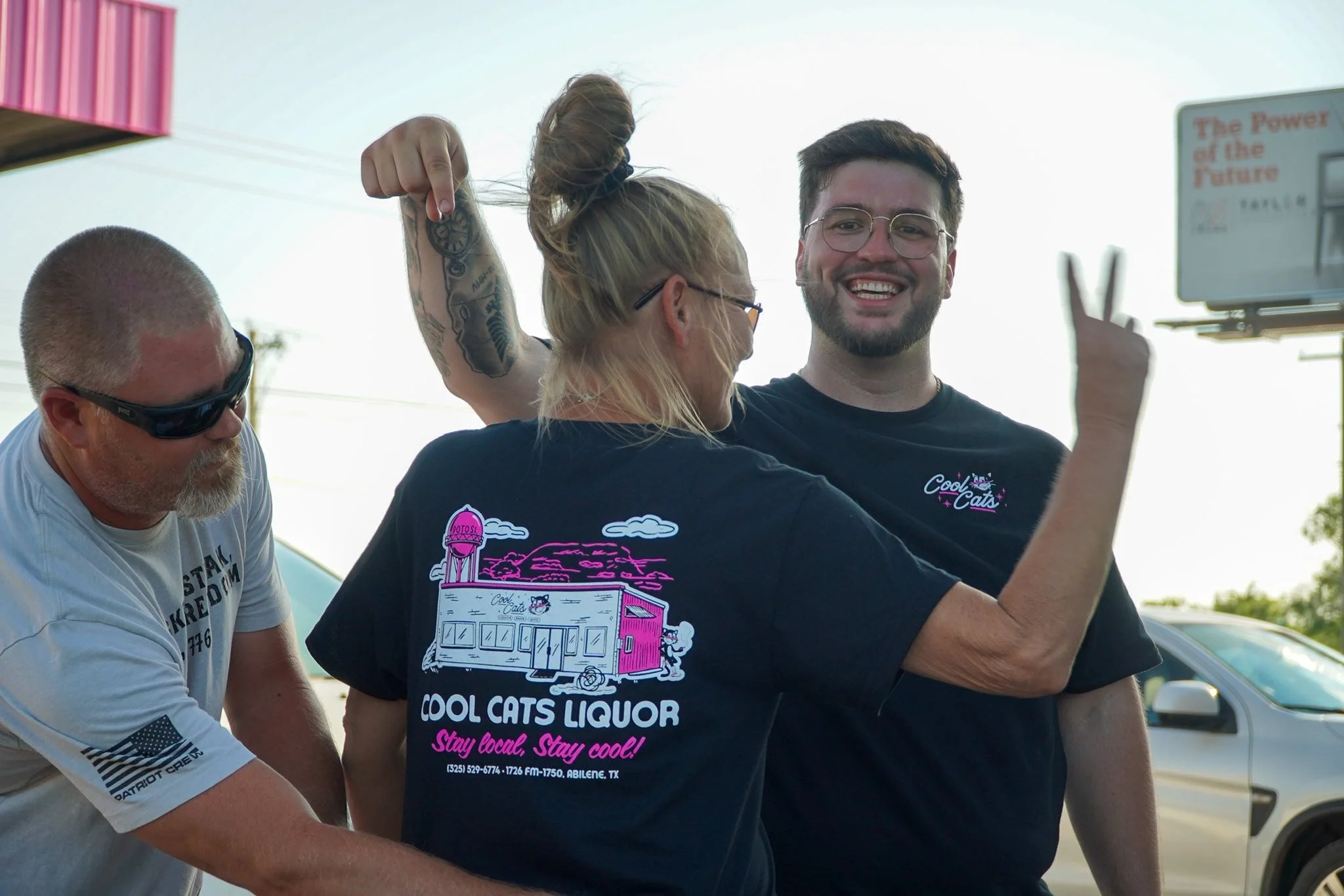 Three friends smiling and joking outdoors, one man with glasses and tattoos showing a peace sign, with two women wearing black T-shirts for Cool Cats Liquor, one woman with glasses and blonde hair in a bun, and the other with sunglasses and a beard, 