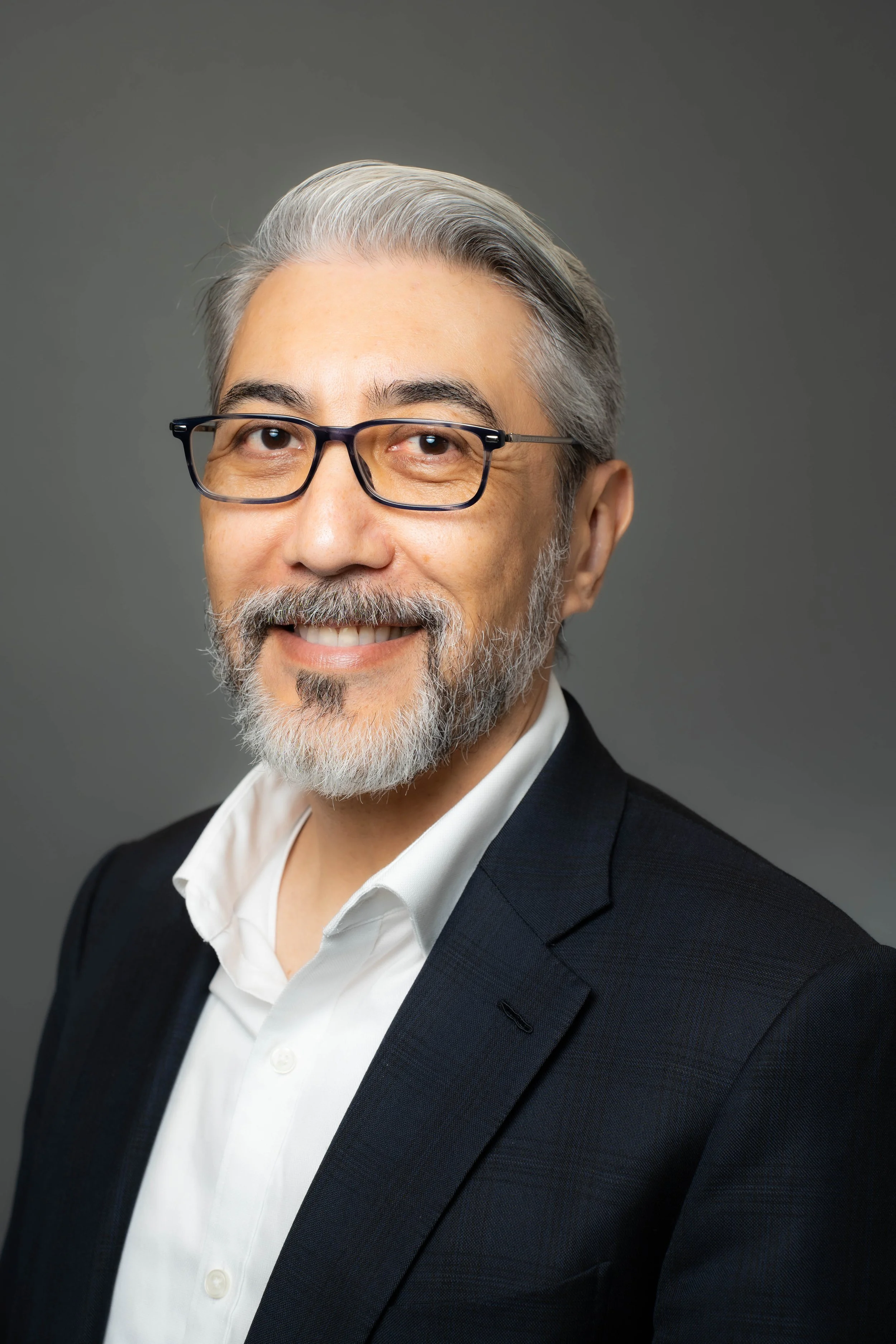 Headshot of a middle-aged man with gray hair, glasses, and a beard, wearing a white shirt and a black blazer, smiling against a gray background.
