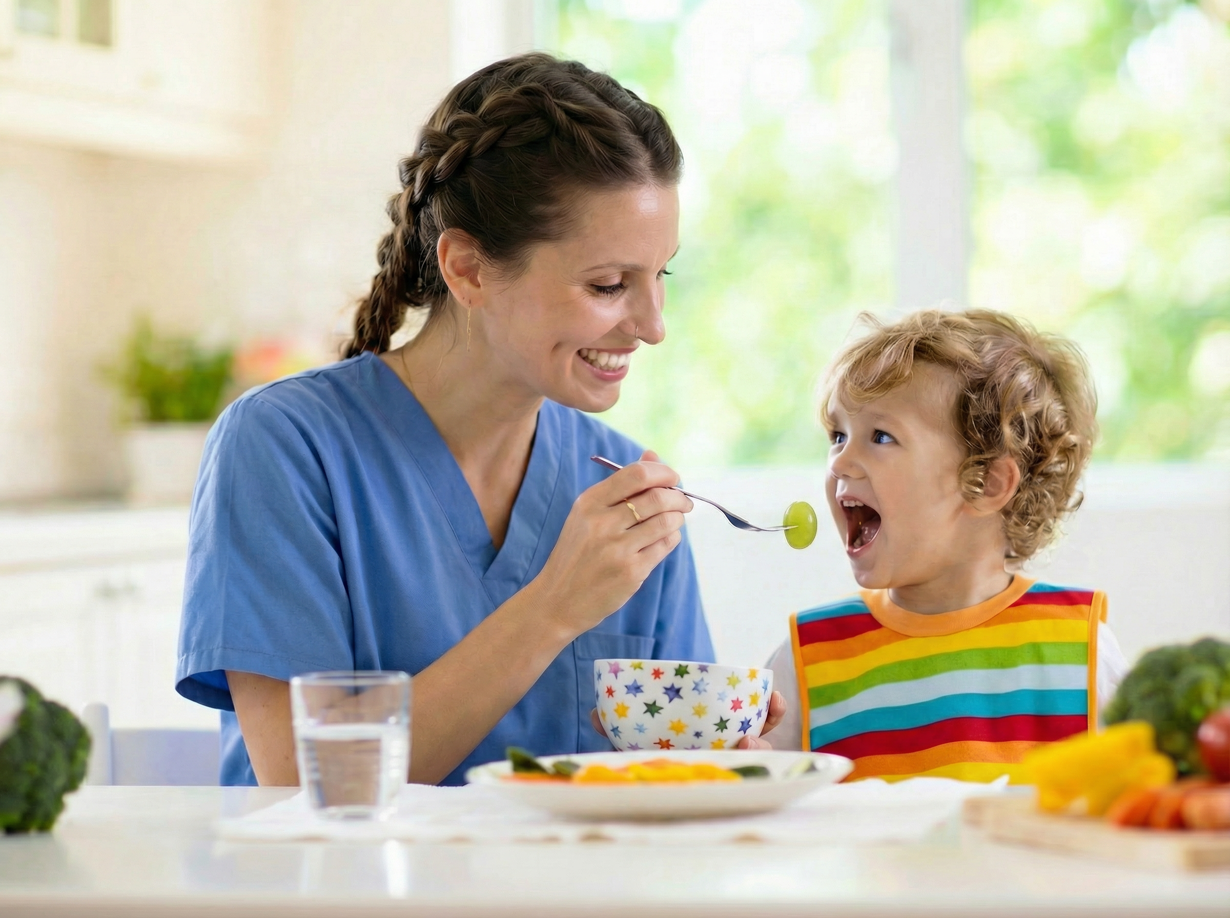 Caregiver feeds a toddler during a pediatric feeding therapy session in a bright kitchen setting.