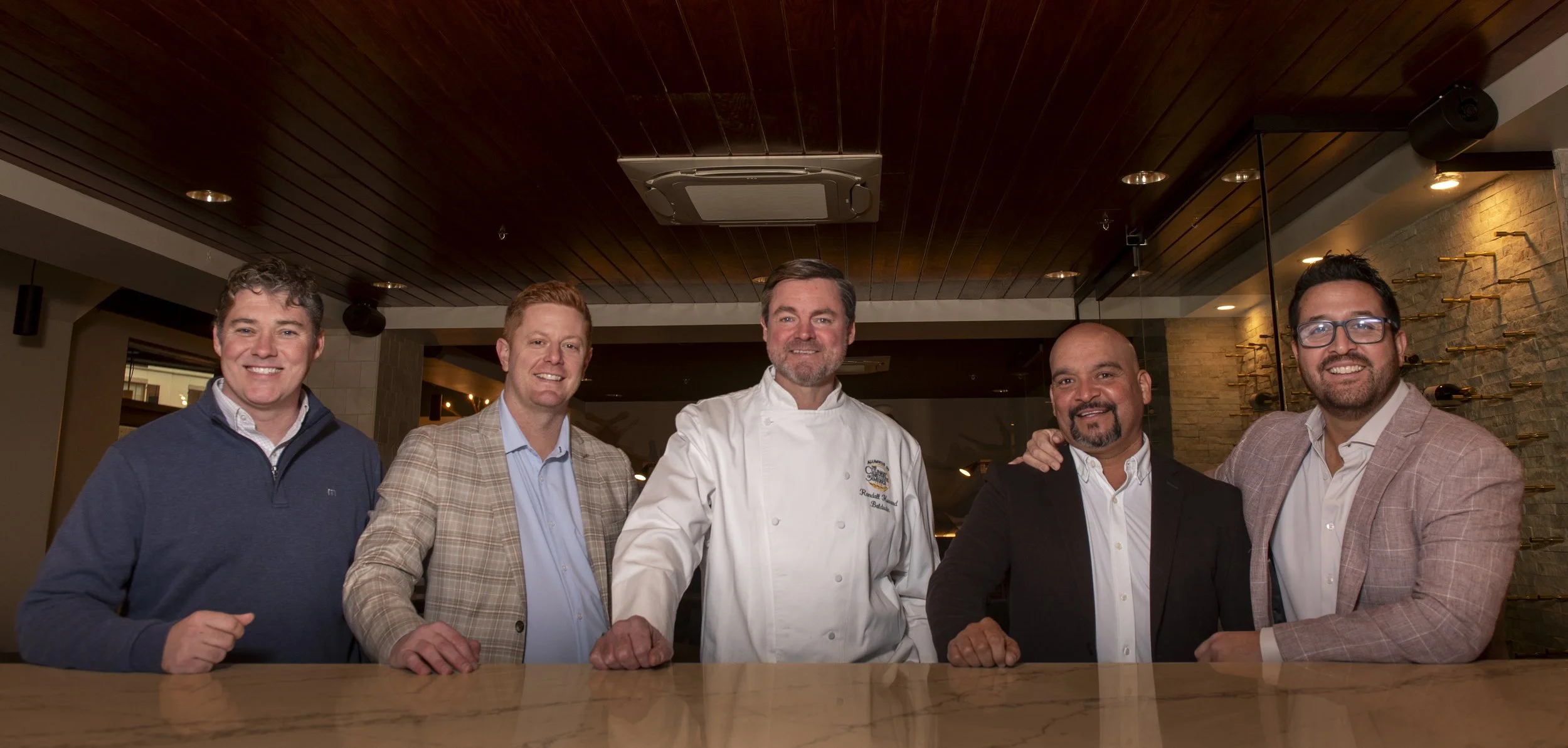 Group of five men standing behind a bar counter, smiling, including a chef in the middle, in a restaurant or bar setting.