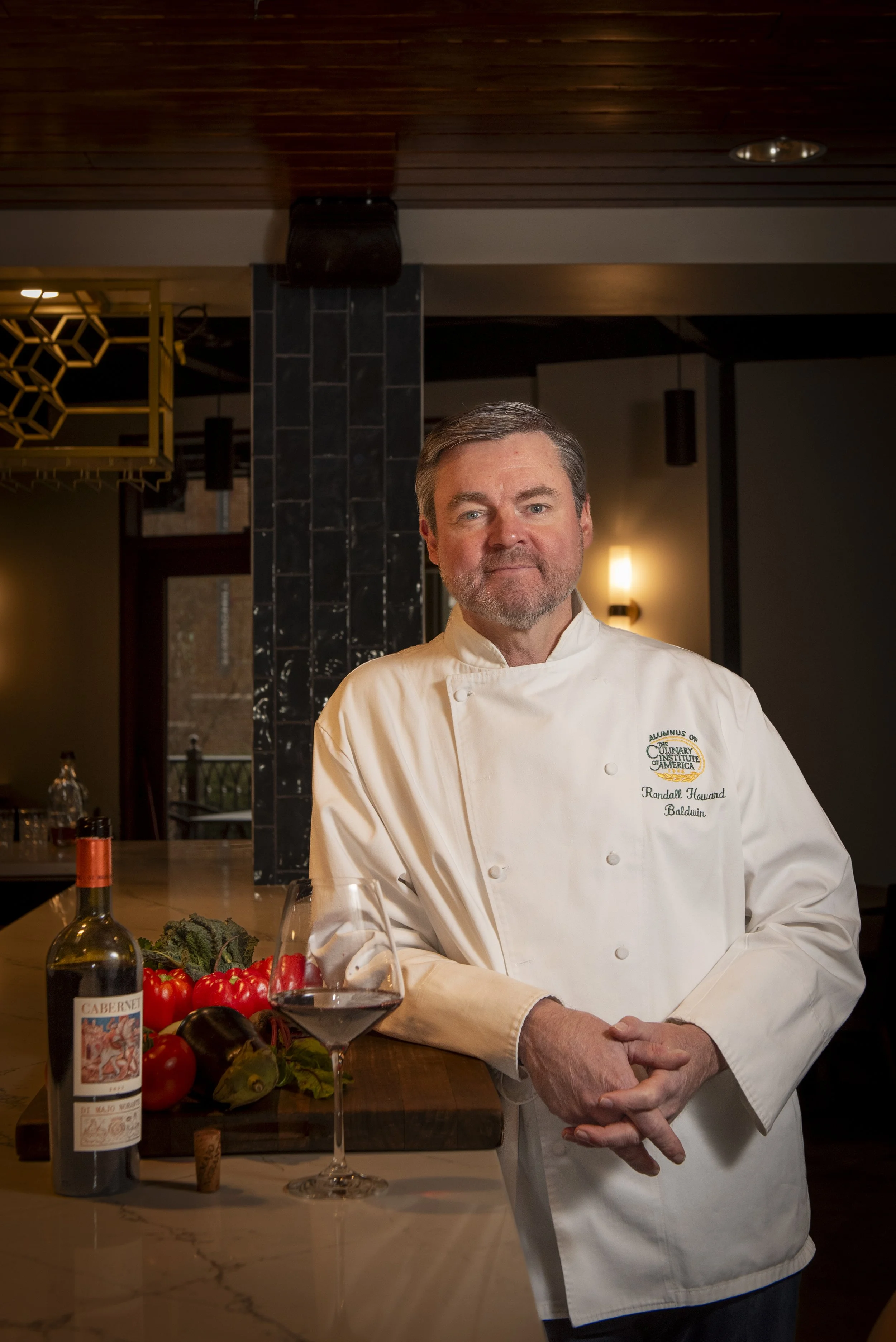 A male chef in a white chef's coat standing in a modern kitchen, with a wine glass of red wine, a bottle of red wine, and fresh vegetables on the counter.