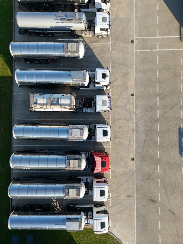 Top-down aerial view of a parking lot with ten semi-trucks parked in designated spaces. The trucks are white, silver, and red, with some having large cargo trailers attached.