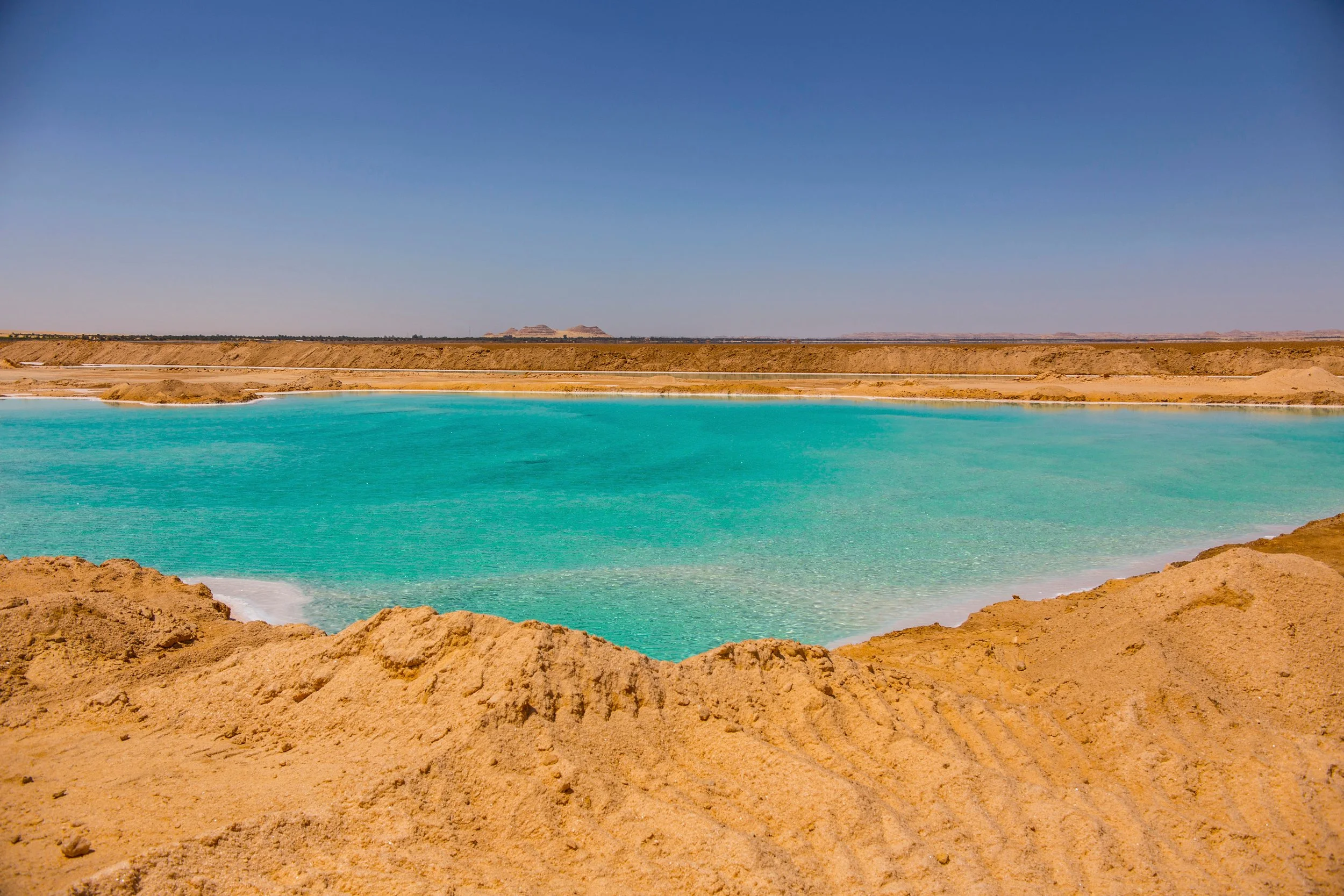 Peaceful desert landscape with a turquoise lake, sandy shores, and flat distant horizon under clear blue sky.