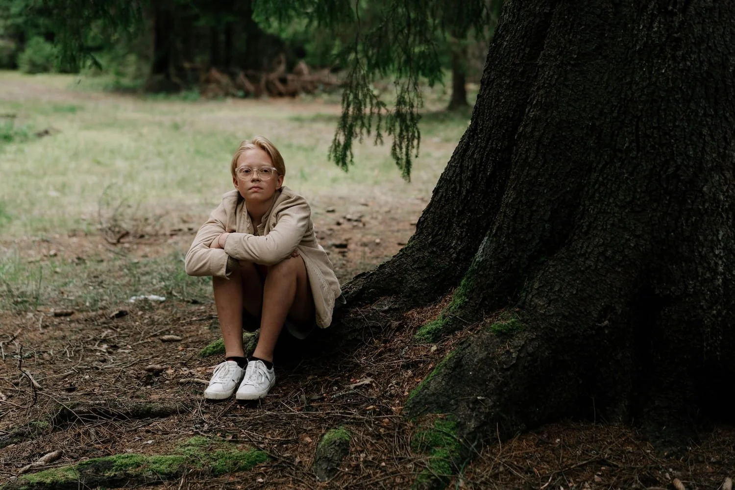 Young person with glasses sitting thoughtfully alone under tree in park