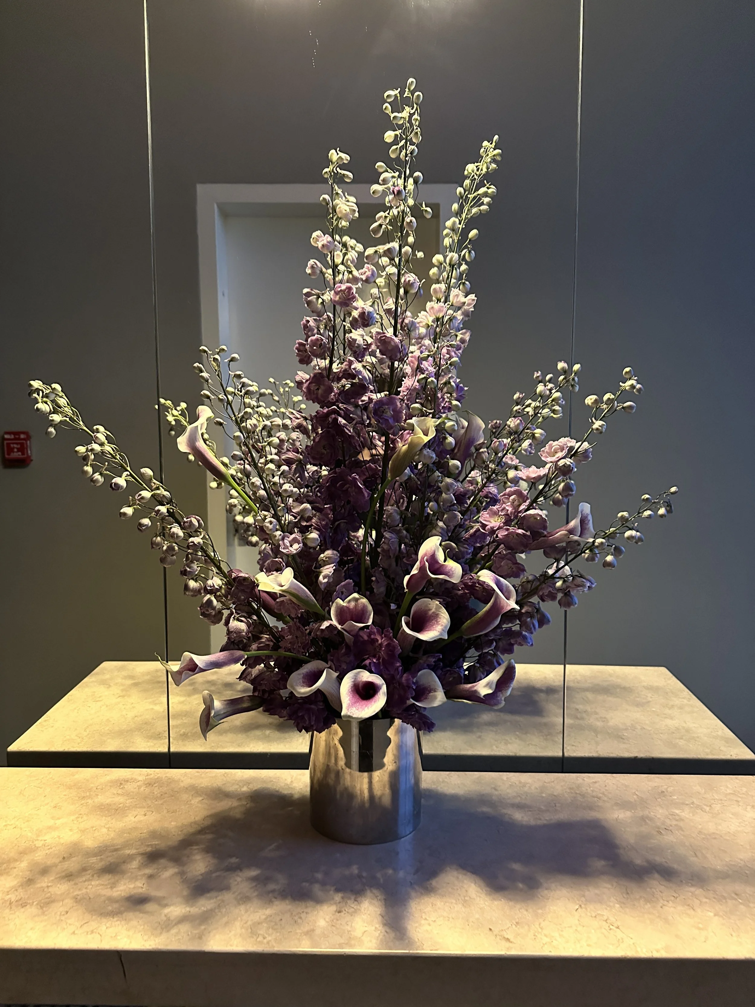 A floral arrangement with pink calla lilies, purple and pink roses, and sprigs of small white flowers in a metallic vase on a beige surface, against a mirrored background.