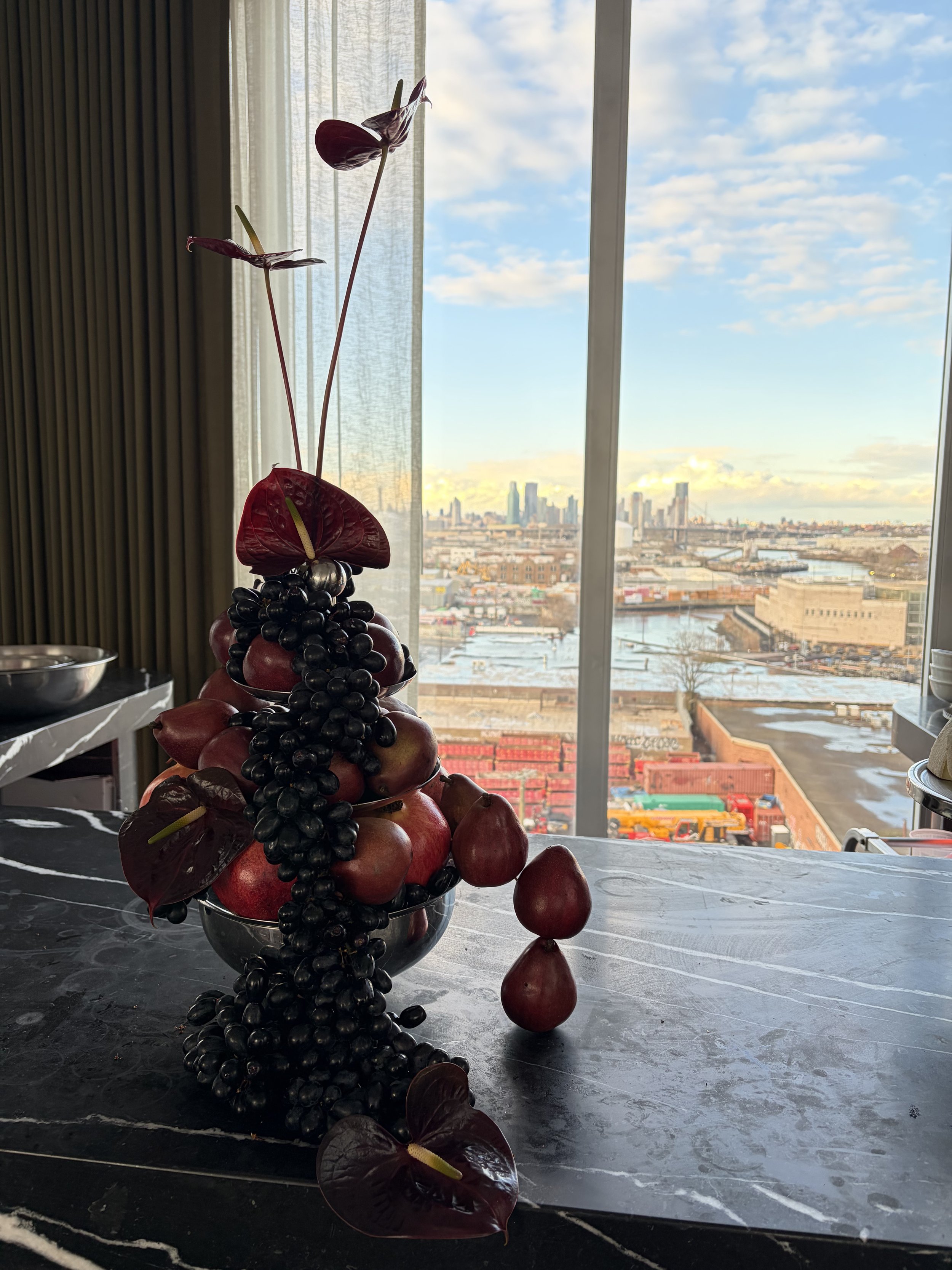 A sculpture made of assorted fruits including apples, grapes, and calla lilies, placed on a dark marble table near large windows with a cityscape view in the background.