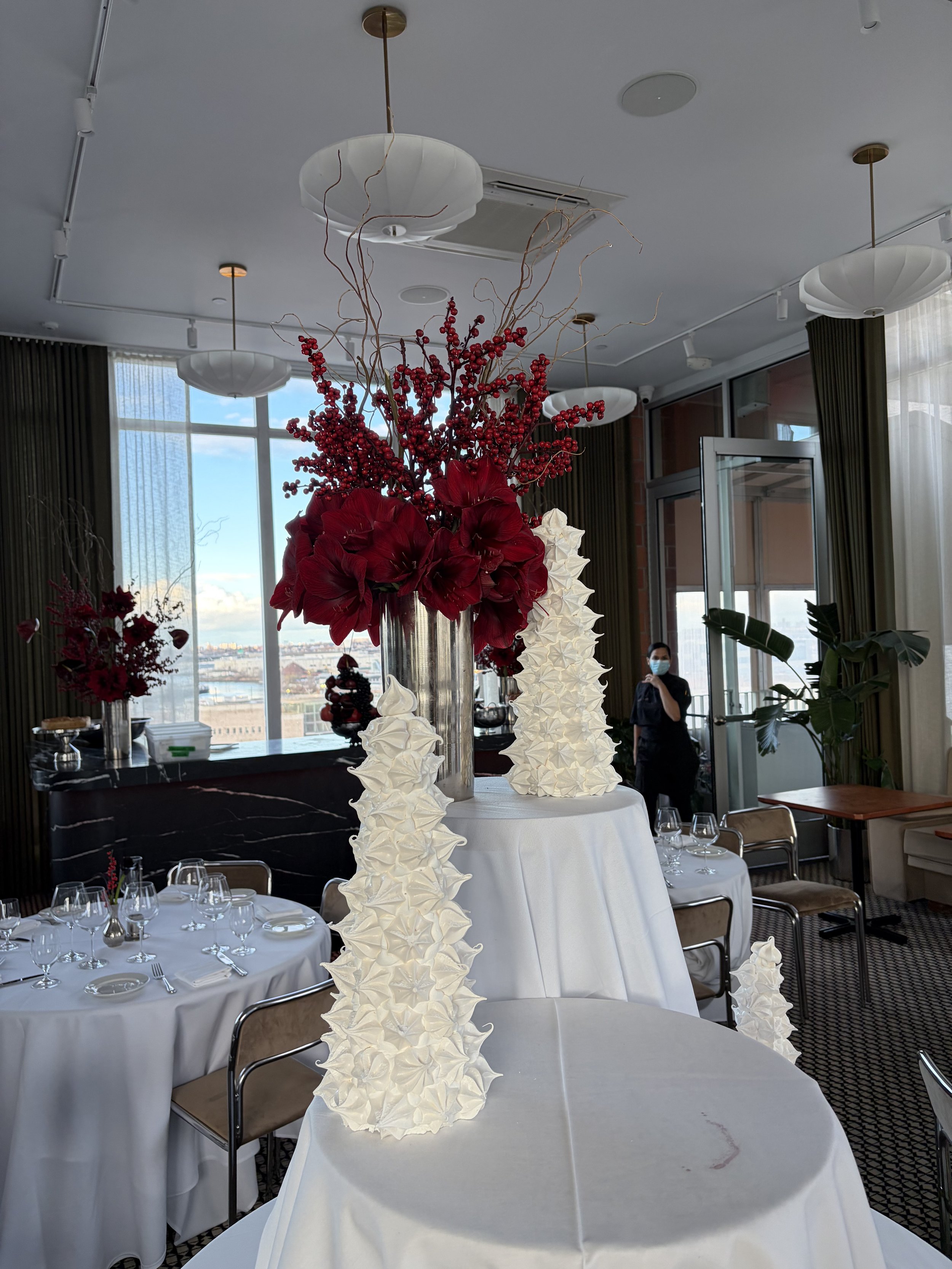Elegant event table with tall floral centerpieces featuring red and white flowers and branches, set in a bright room with large windows, white tablecloths, and modern decor.