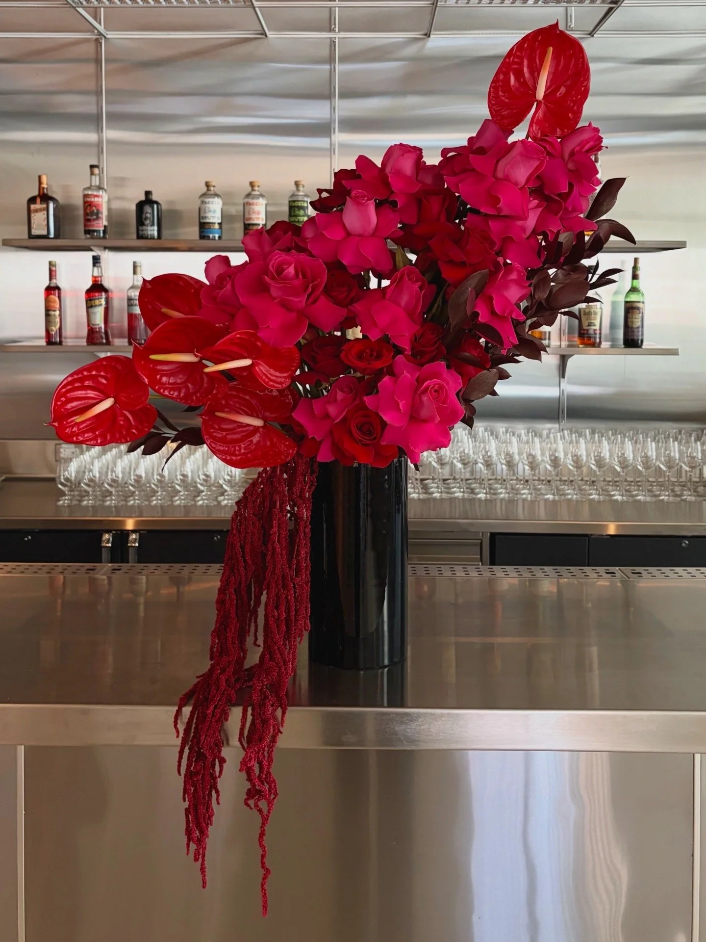 A tall black vase filled with a vibrant arrangement of pink, red, and burgundy flowers, including roses, calla lilies, and anthuriums, with hanging red amaranth, set on a bar counter with a mirrored bar backdrop in a modern setting.