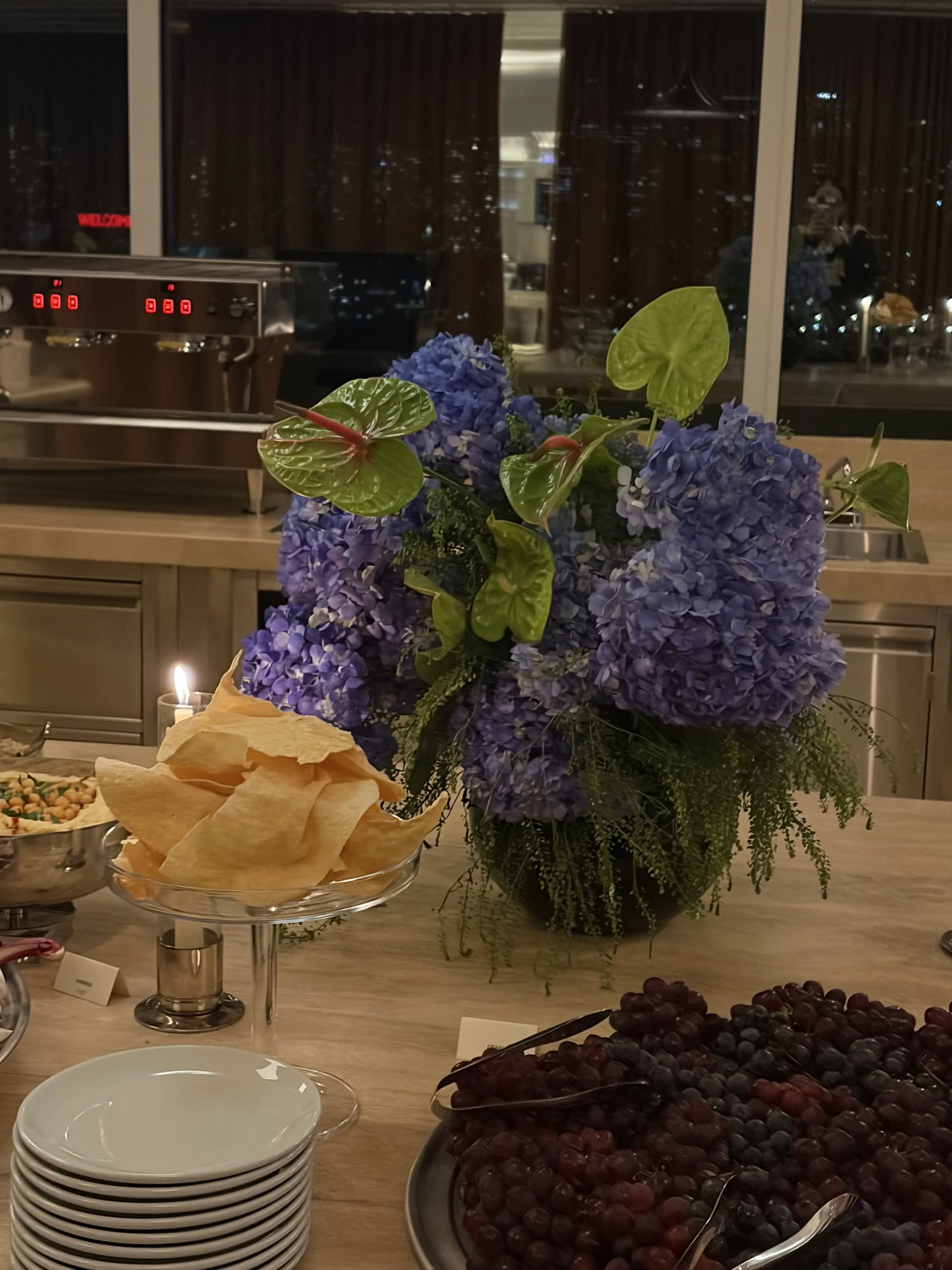 A table with a large blue and green floral arrangement, a stack of small white plates, a bowl of potato chips, and a tray of mixed berries in a modern room with large windows at night.